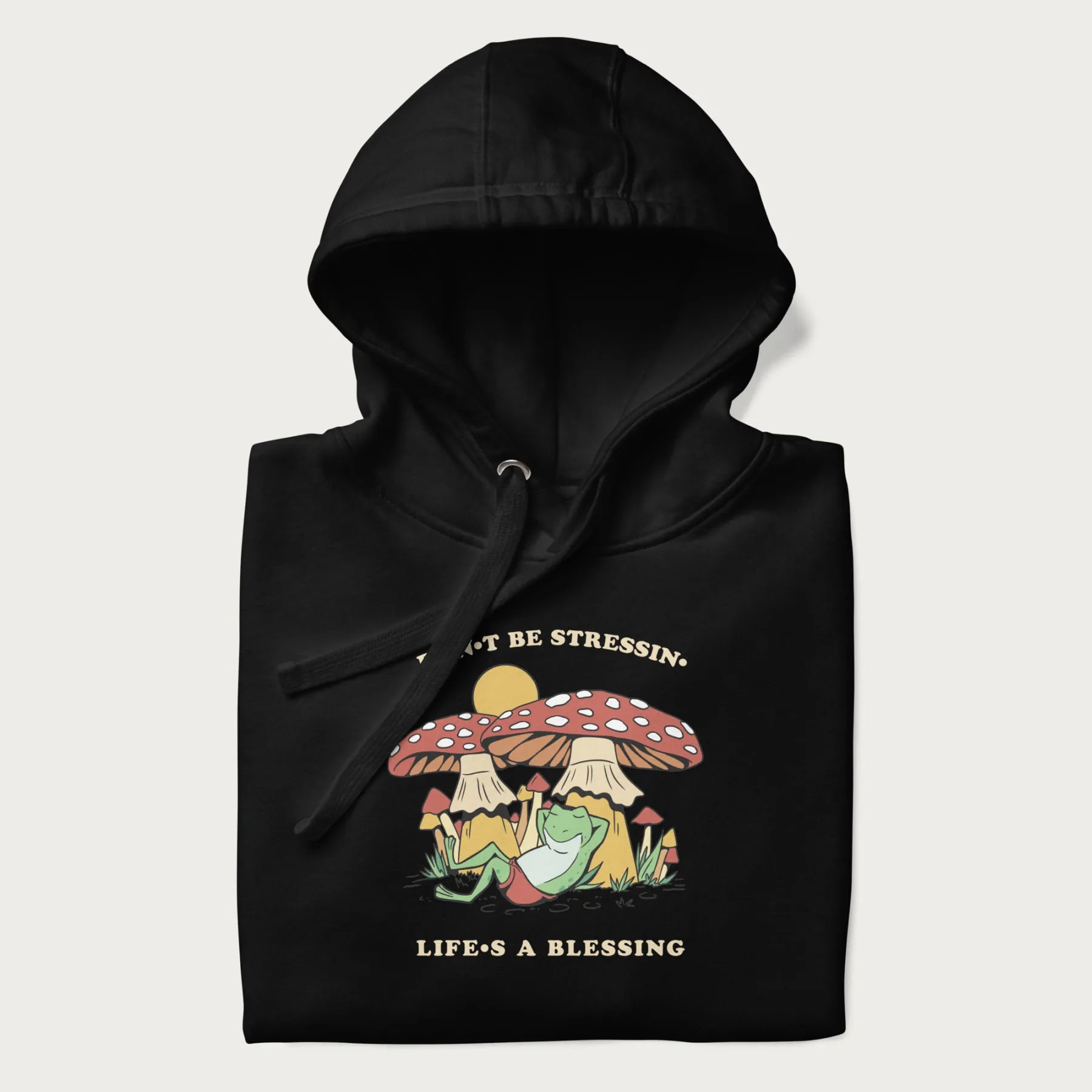 Folded black hoodie with a graphic of a frog lounging under mushrooms and the phrases 'Don't Be Stressin' Life's a Blessing'.