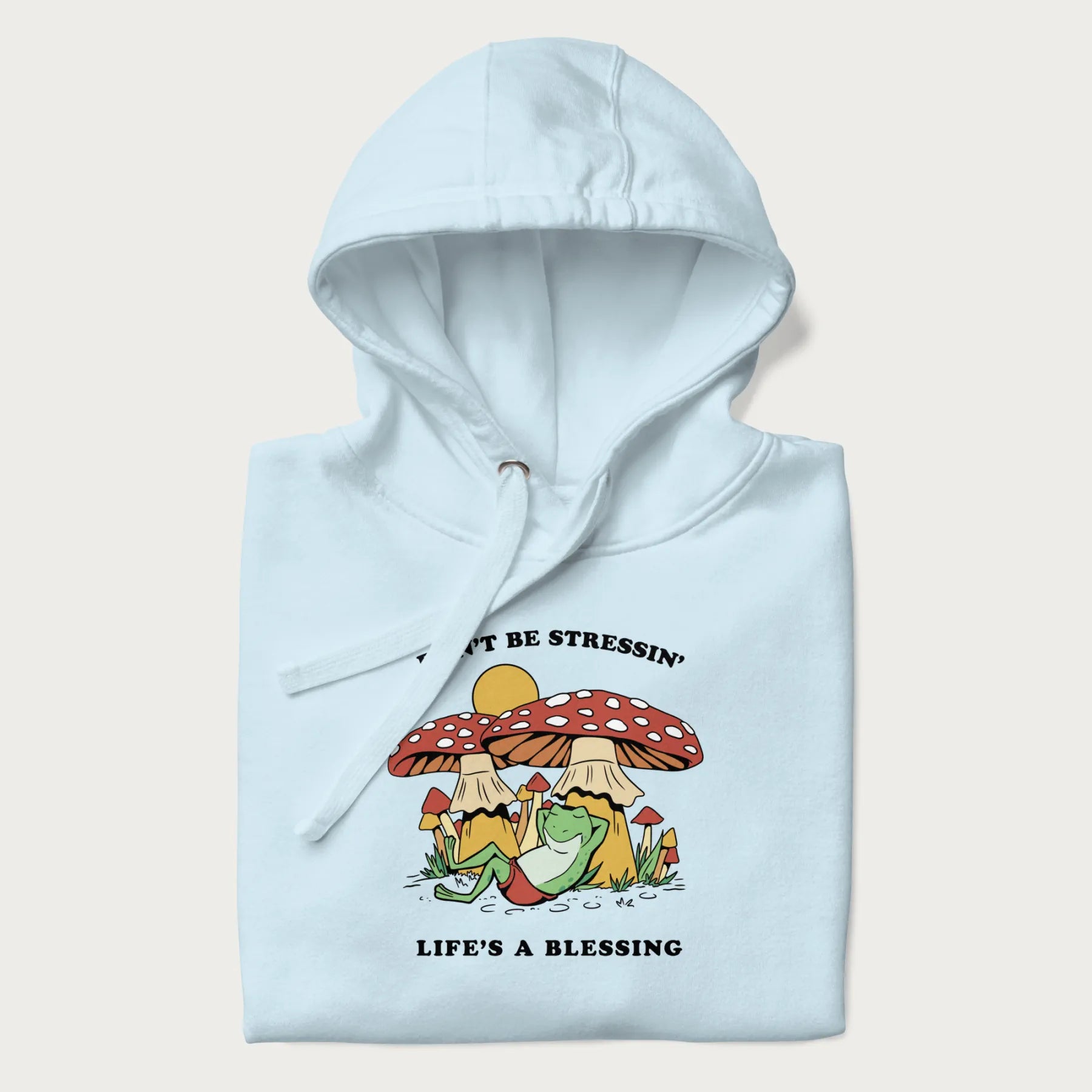Folded light blue hoodie with a graphic of a frog lounging under mushrooms and the phrases 'Don't Be Stressin' Life's a Blessing'.