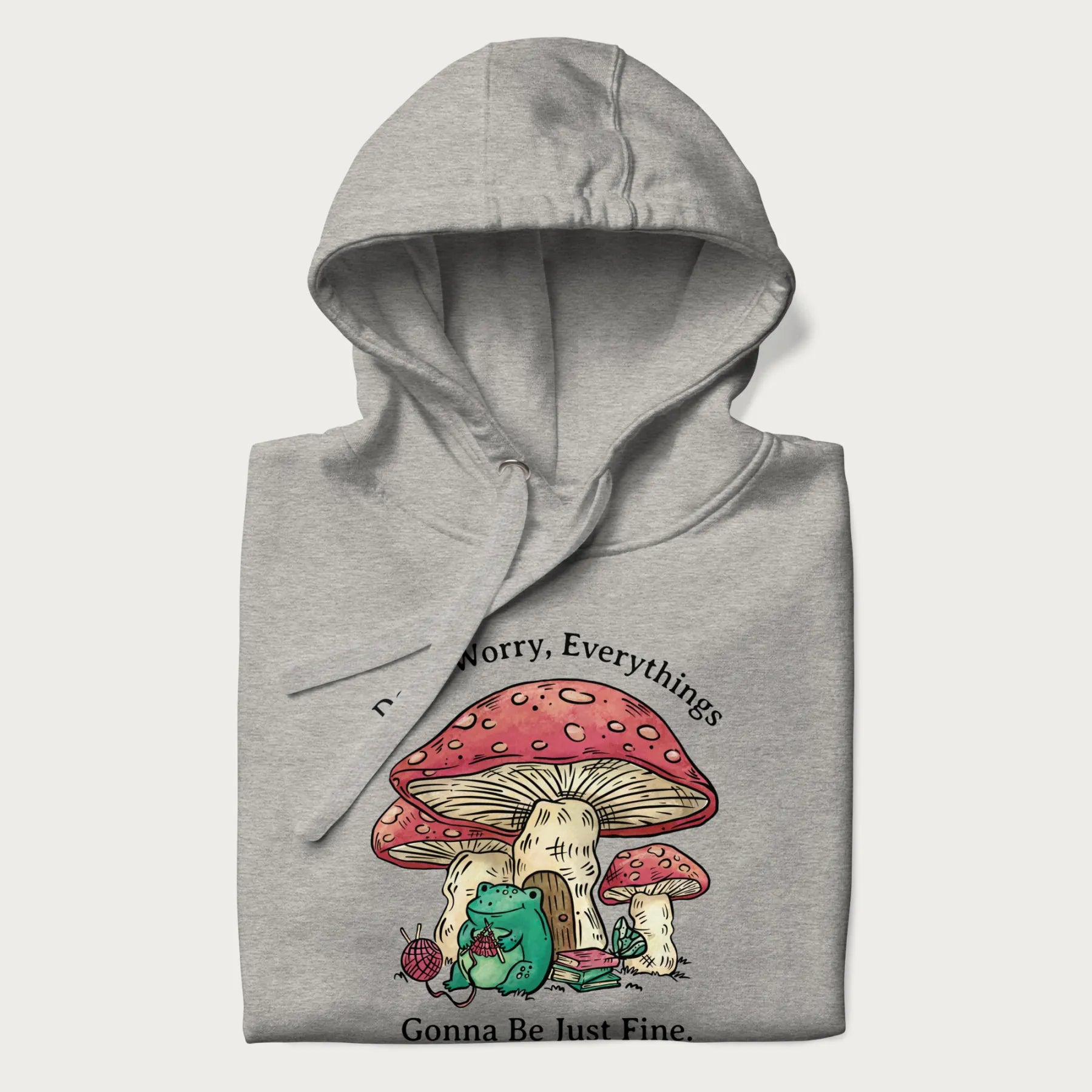 Folded light grey hoodie with a graphic of a frog knitting under a large mushroom with the text 'Don't Worry, Everything's Gonna Be Just Fine'.