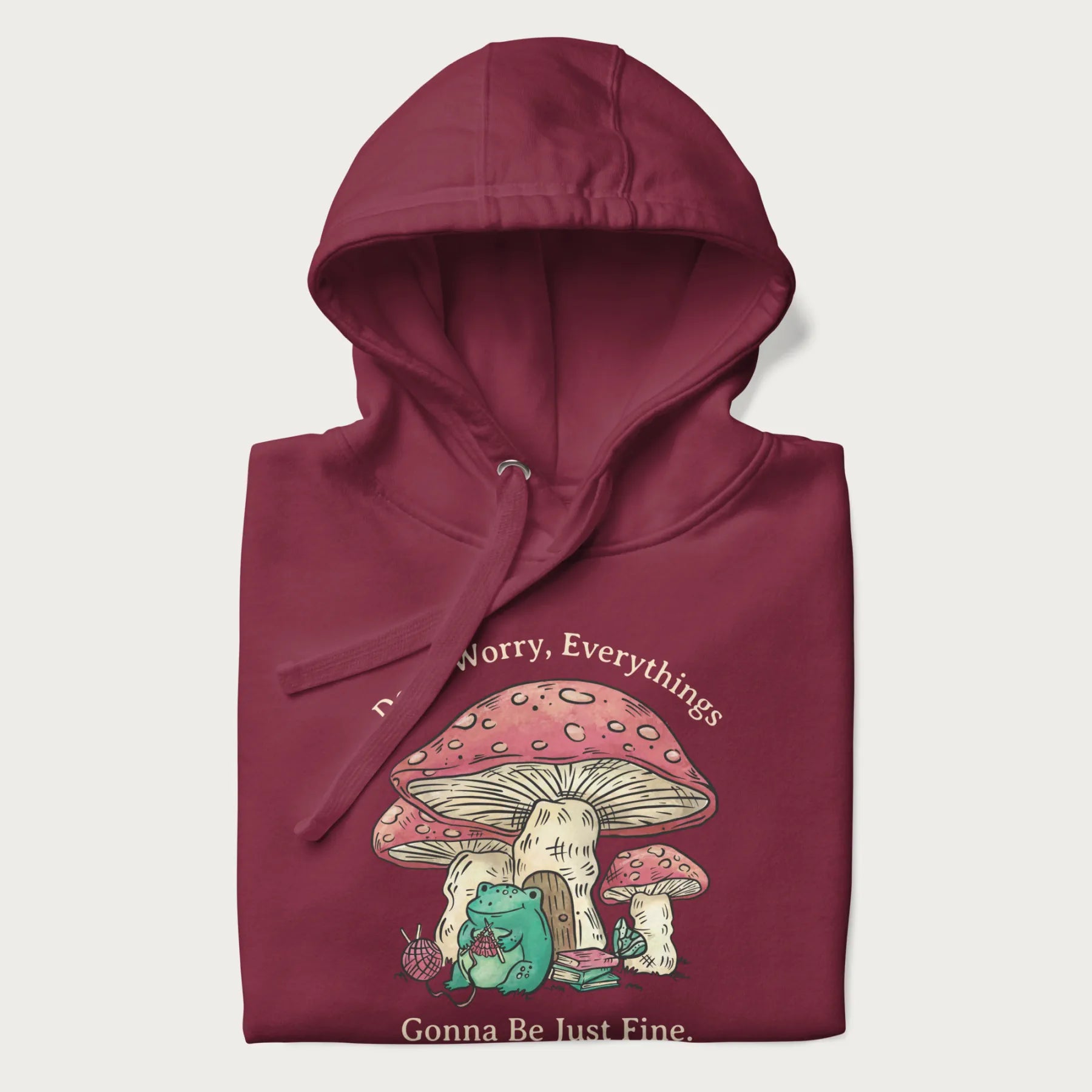 Folded maroon hoodie with a graphic of a frog knitting under a large mushroom with the text 'Don't Worry, Everything's Gonna Be Just Fine'.
