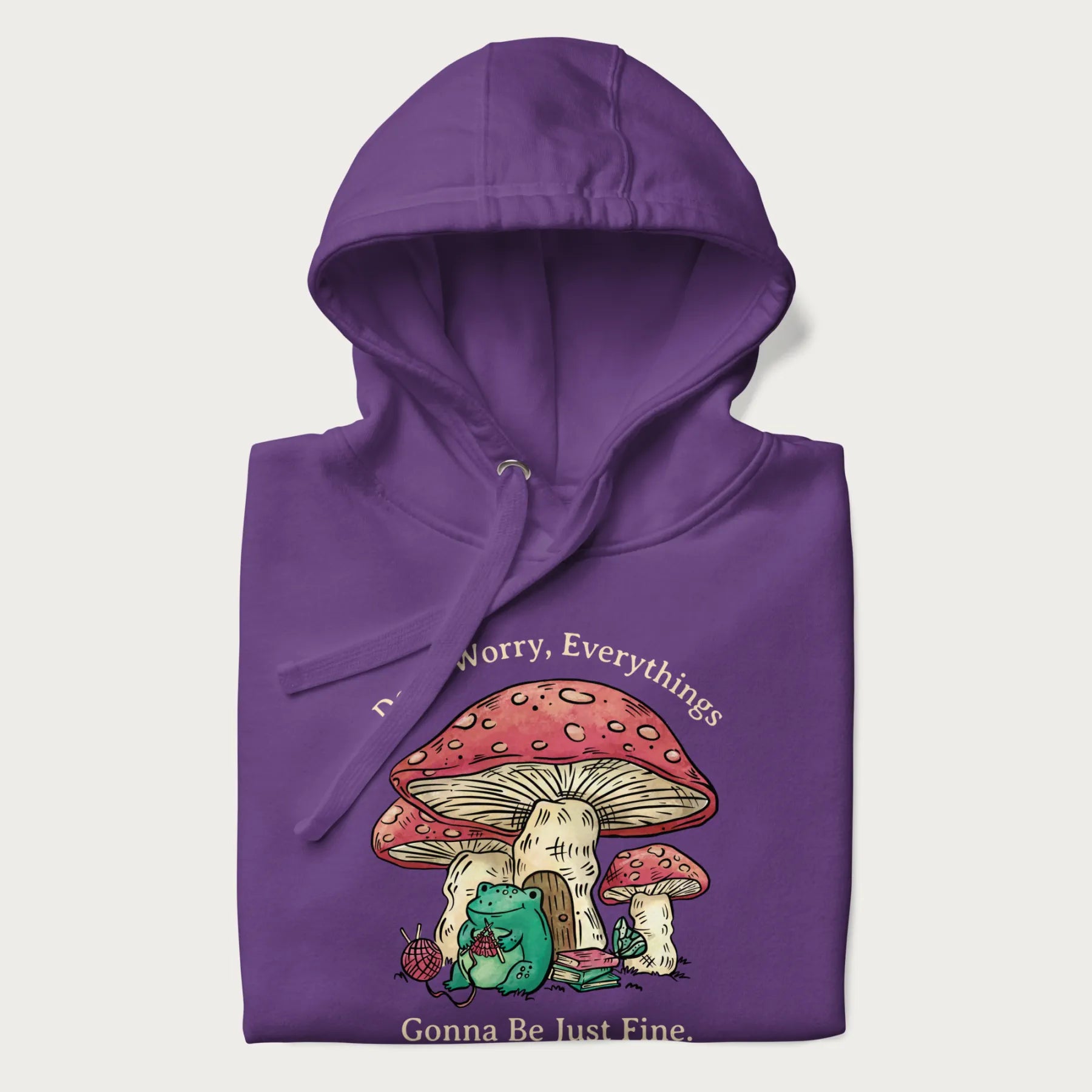 Folded purple hoodie with a graphic of a frog knitting under a large mushroom with the text 'Don't Worry, Everything's Gonna Be Just Fine'.