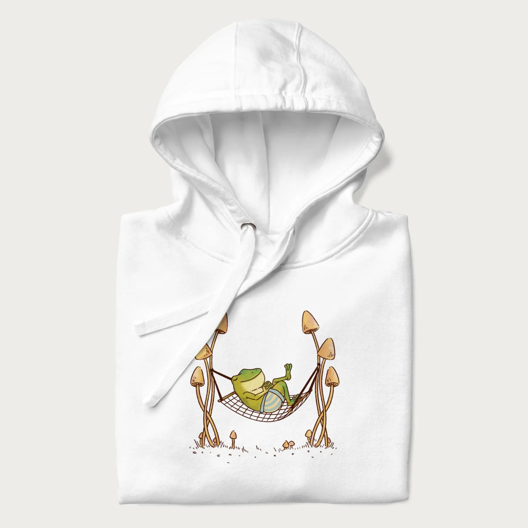 Folded white hoodie with a graphic of a frog lounging in a hammock between tall mushrooms.