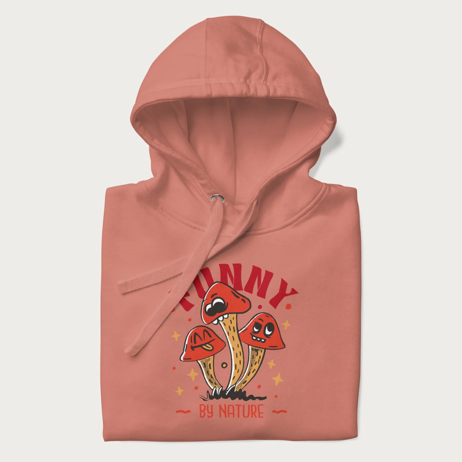 Folded light pink hoodie featuring a playful graphic of three mushrooms with expressive faces and the text "Funny by Nature."