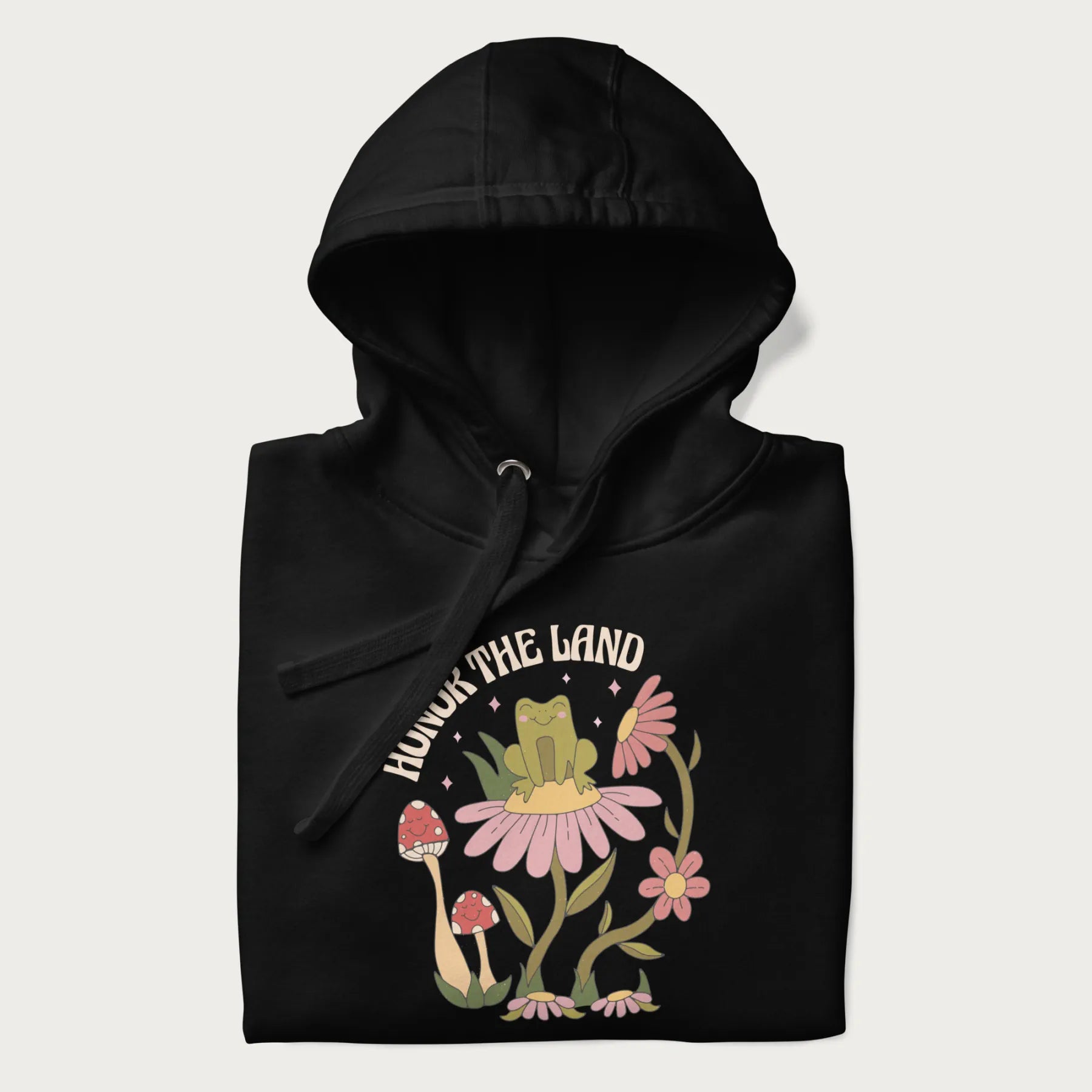 Folded black hoodie with a graphic of a frog sitting on a flower, surrounded by mushrooms and flowers, with the text 'Honor the Land'.