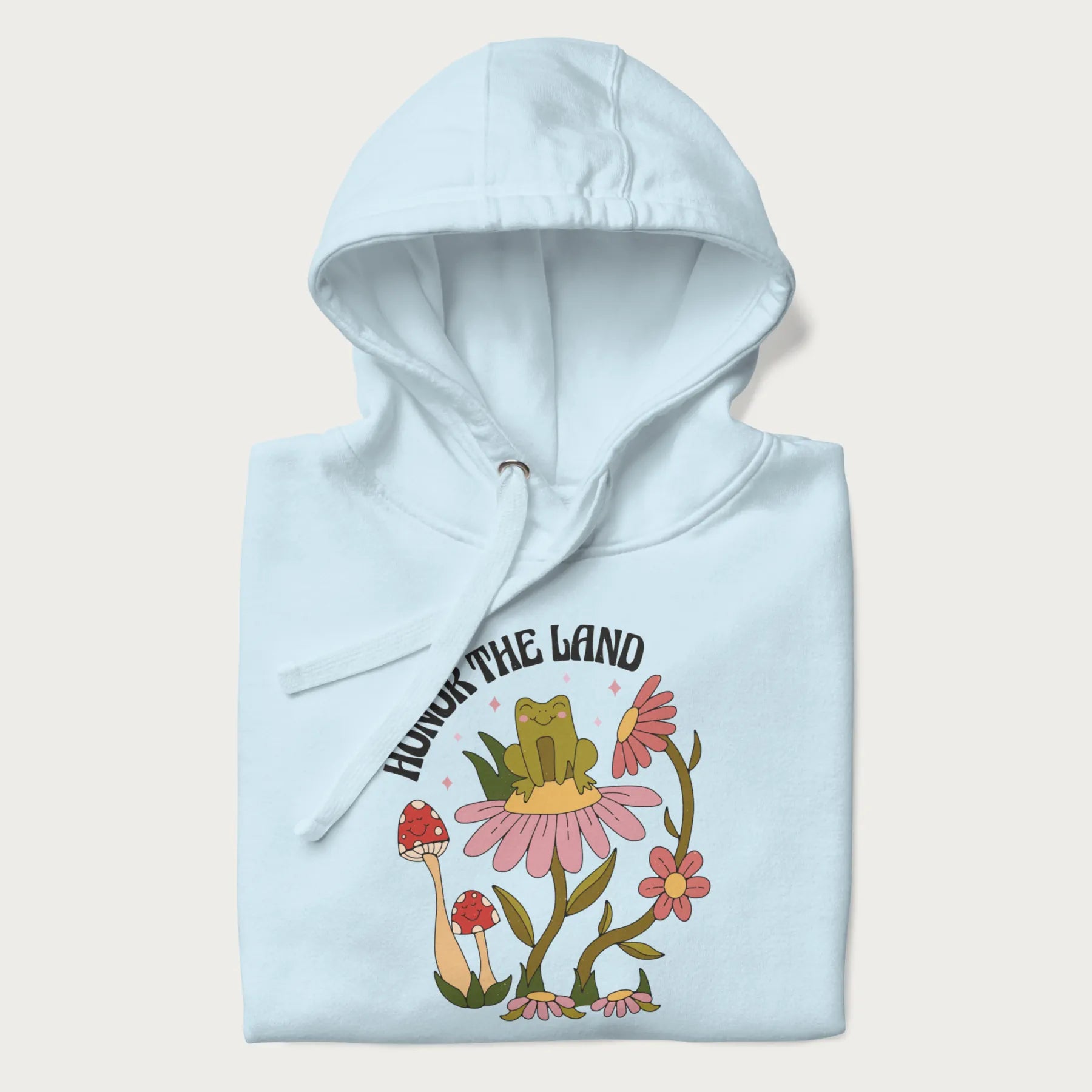 Folded light blue hoodie with a graphic of a frog sitting on a flower, surrounded by mushrooms and flowers, with the text 'Honor the Land'.