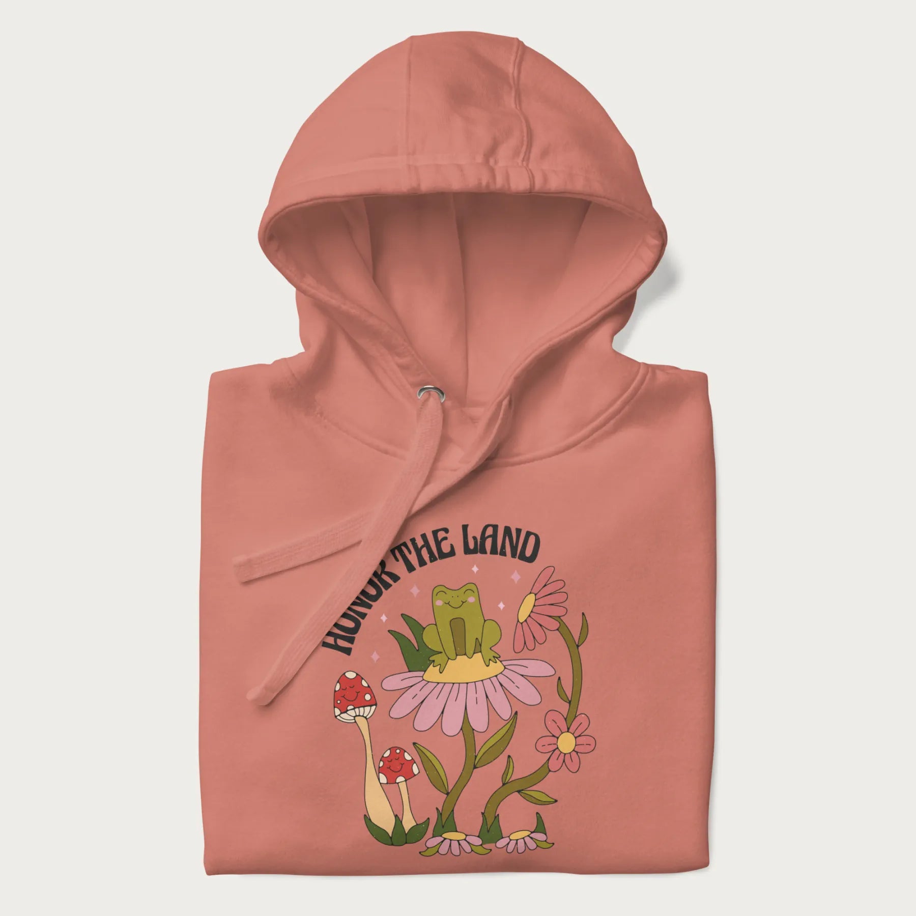 Folded light pink hoodie with a graphic of a frog sitting on a flower, surrounded by mushrooms and flowers, with the text 'Honor the Land'.