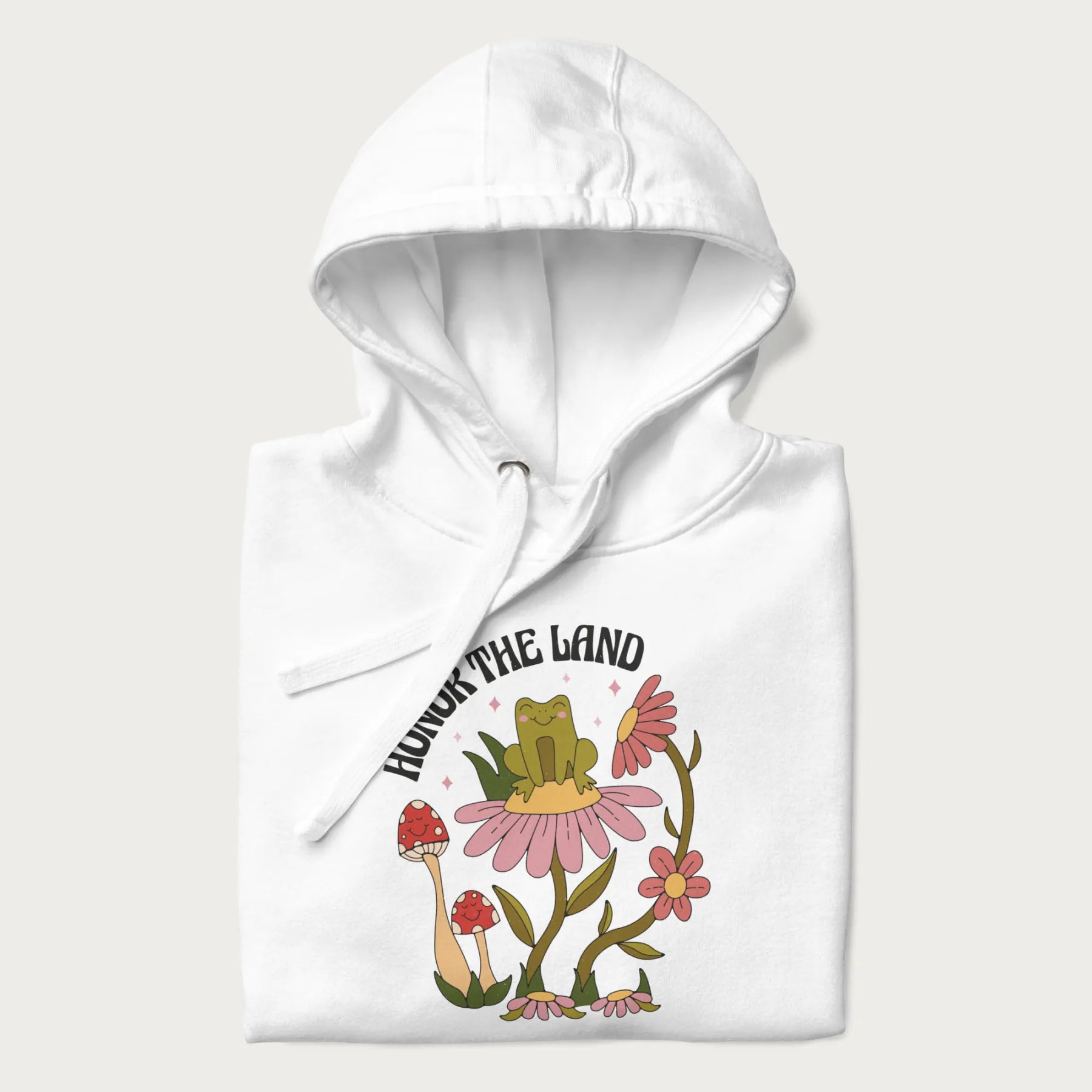 Folded white hoodie with a graphic of a frog sitting on a flower, surrounded by mushrooms and flowers, with the text 'Honor the Land'.