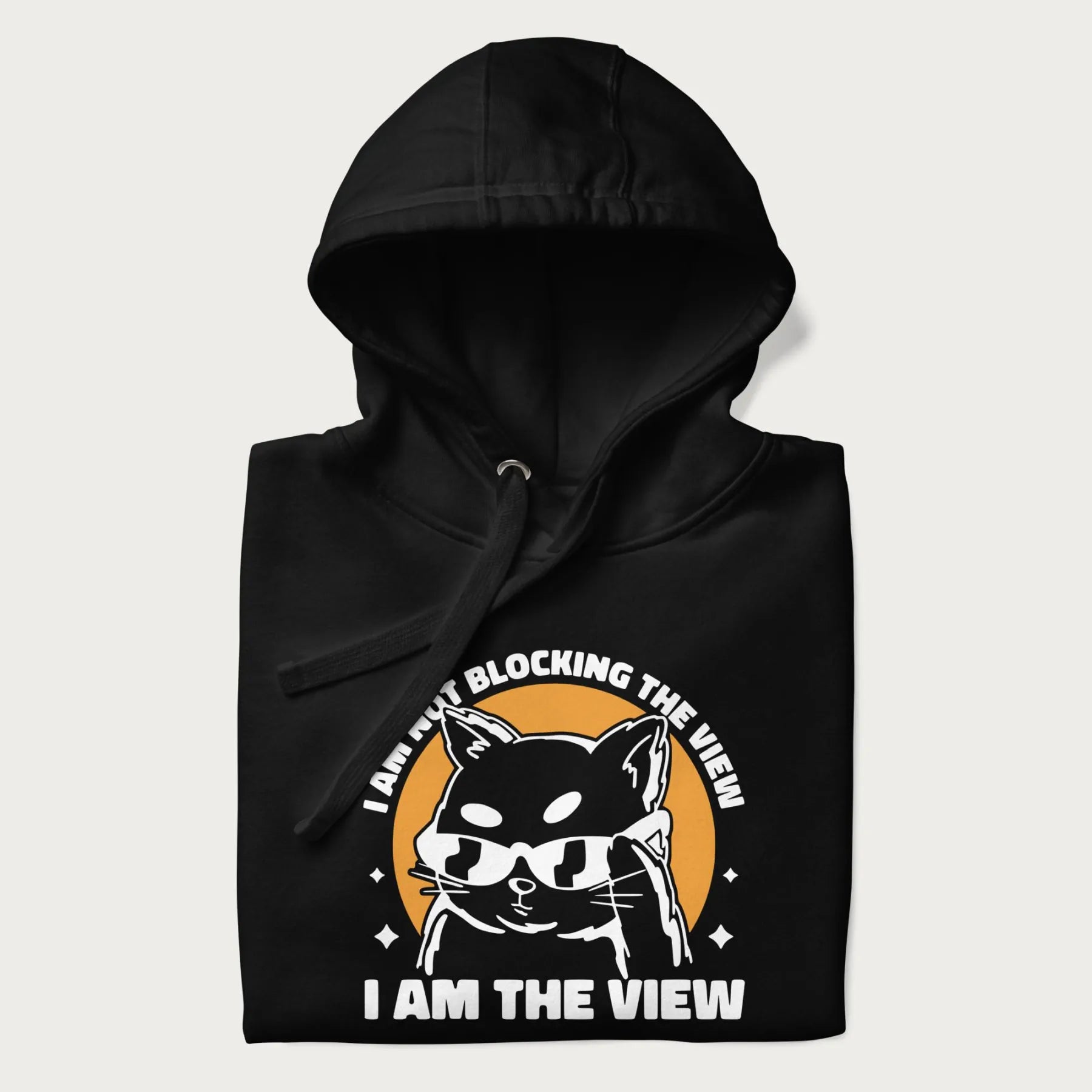 Folded black hoodie with a confident cat wearing sunglasses and the phrase 'I am not blocking the view, I am the view'.