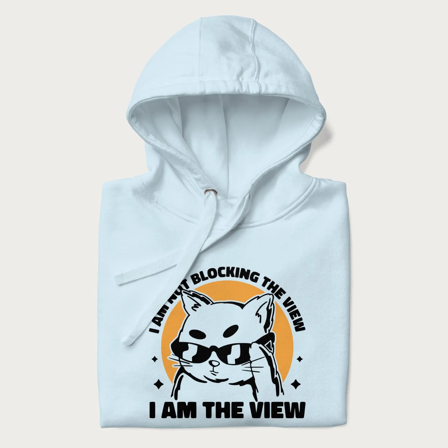 Folded light blue hoodie with a confident cat wearing sunglasses and the phrase 'I am not blocking the view, I am the view'.