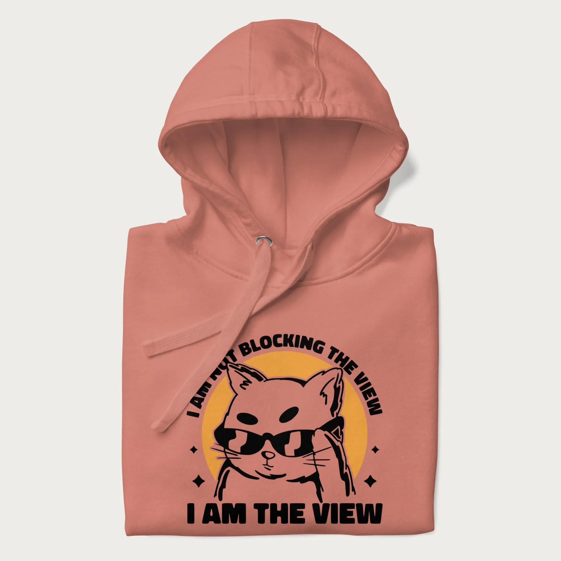 Folded light pink hoodie with a confident cat wearing sunglasses and the phrase 'I am not blocking the view, I am the view'.