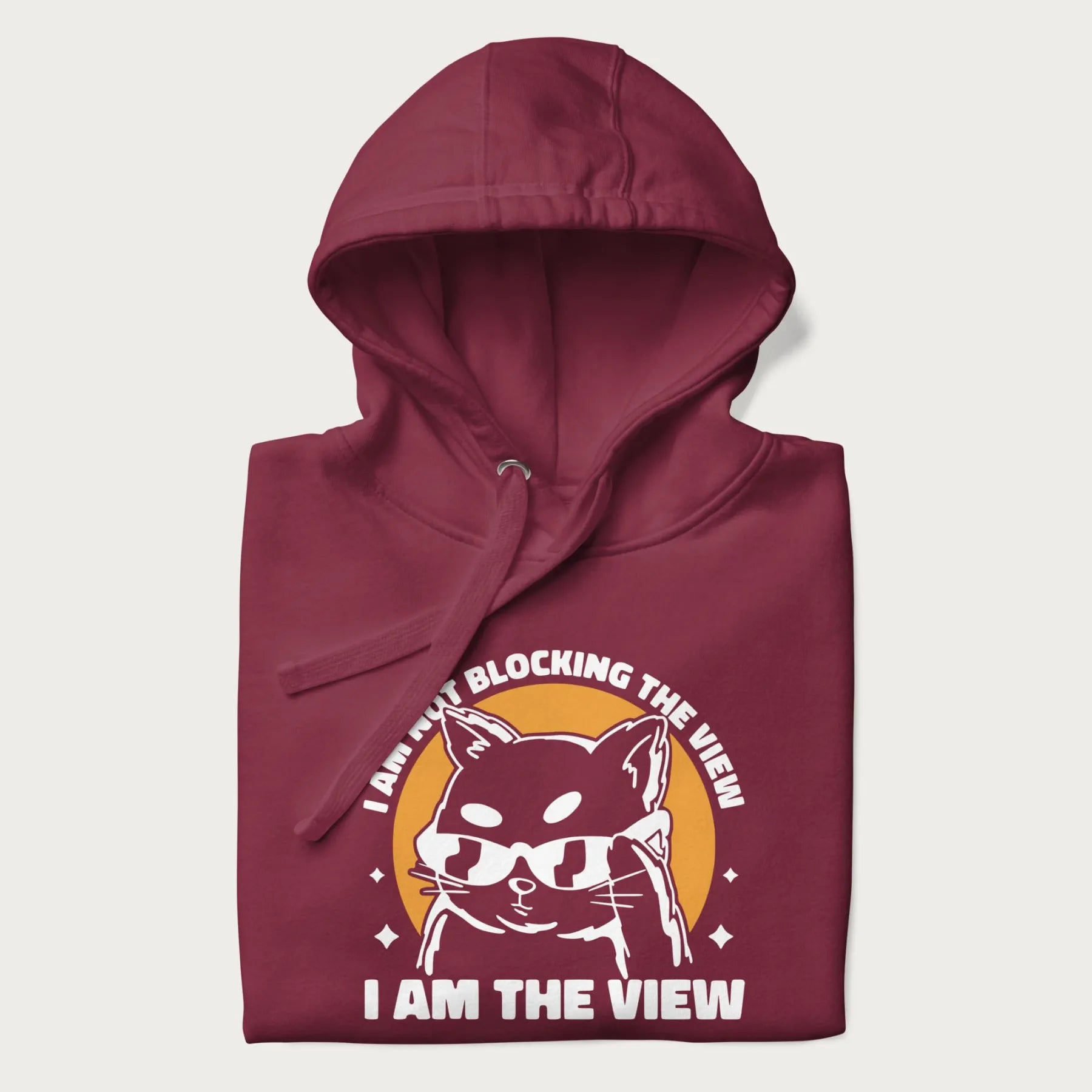 Folded maroon hoodie with a confident cat wearing sunglasses and the phrase 'I am not blocking the view, I am the view'.
