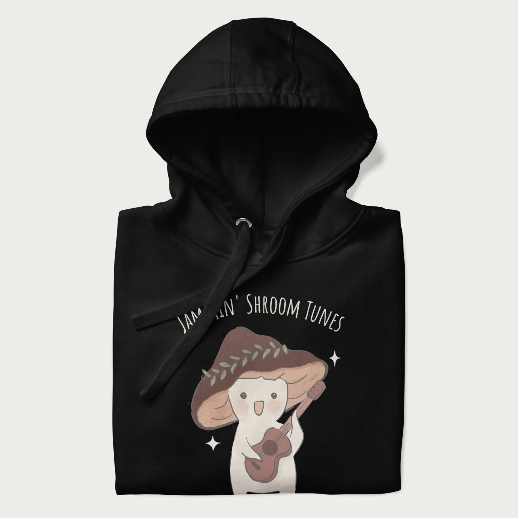 Folded black hoodie with a cute cottagecore mushroom character playing a guitar and the pun 'Jammin' Shroom Tunes' above.