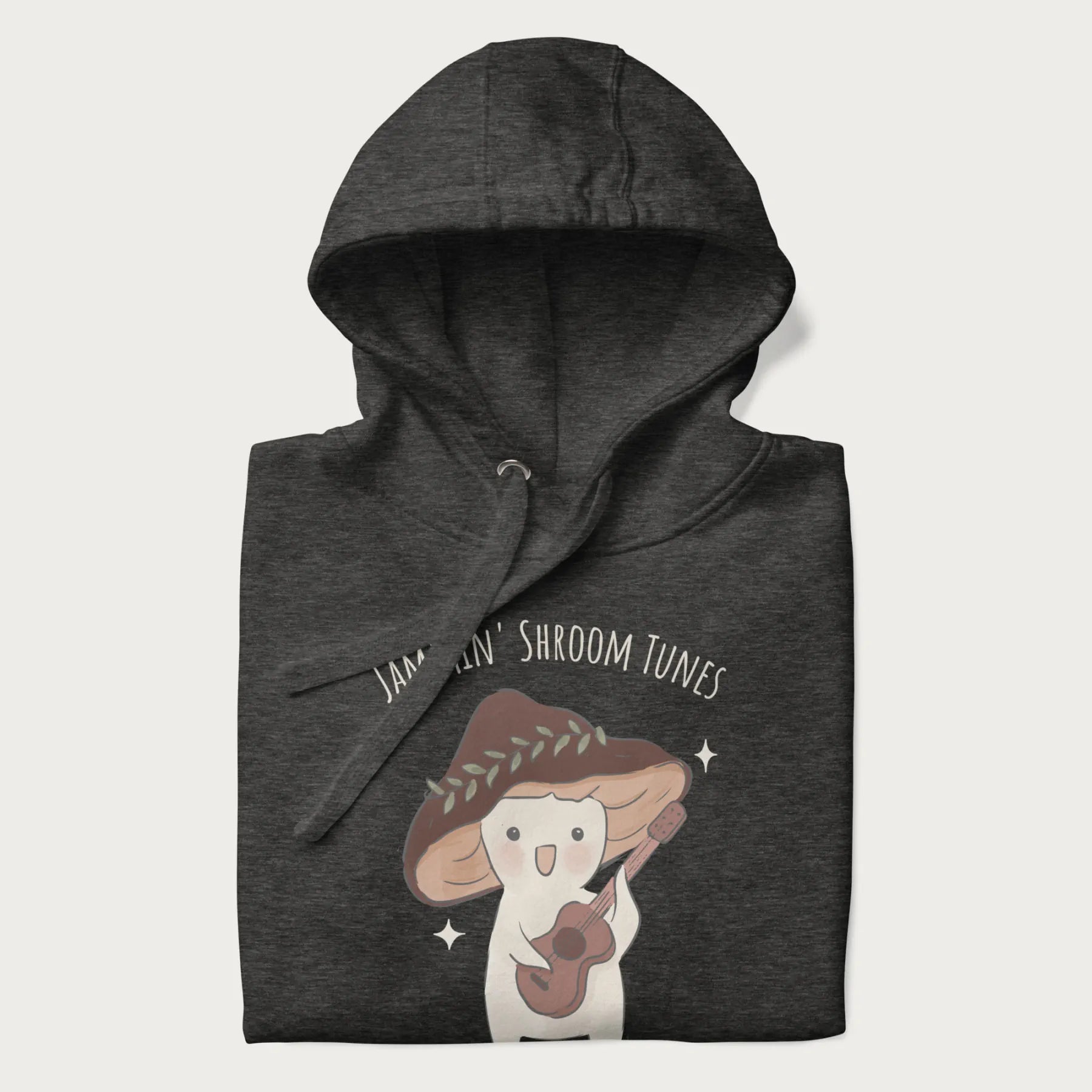 Folded dark grey hoodie with a cute cottagecore mushroom character playing a guitar and the pun 'Jammin' Shroom Tunes' above.