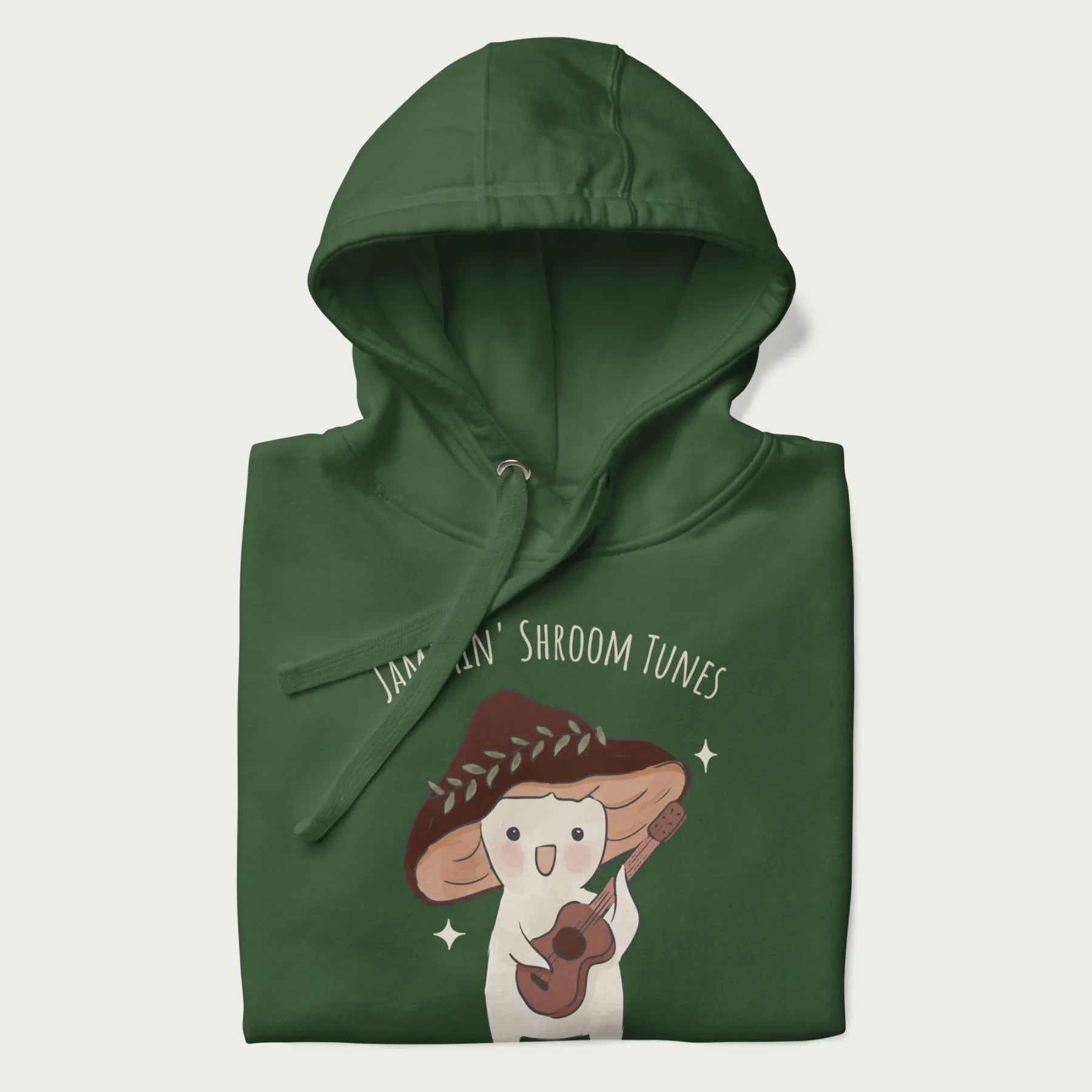 Folded forest green hoodie with a cute cottagecore mushroom character playing a guitar and the pun 'Jammin' Shroom Tunes' above.