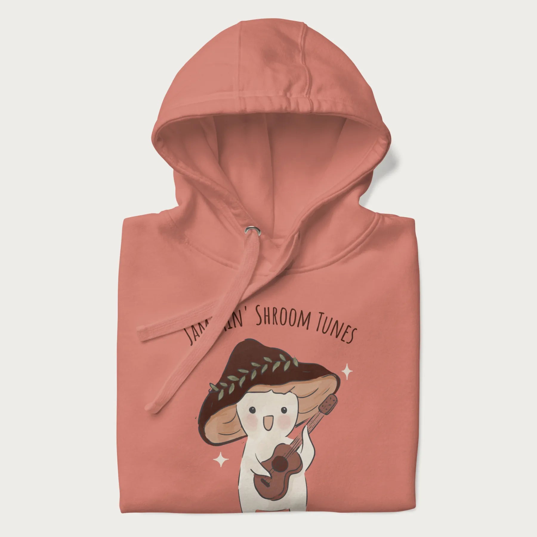 Folded light pink hoodie with a cute cottagecore mushroom character playing a guitar and the pun 'Jammin' Shroom Tunes' above.