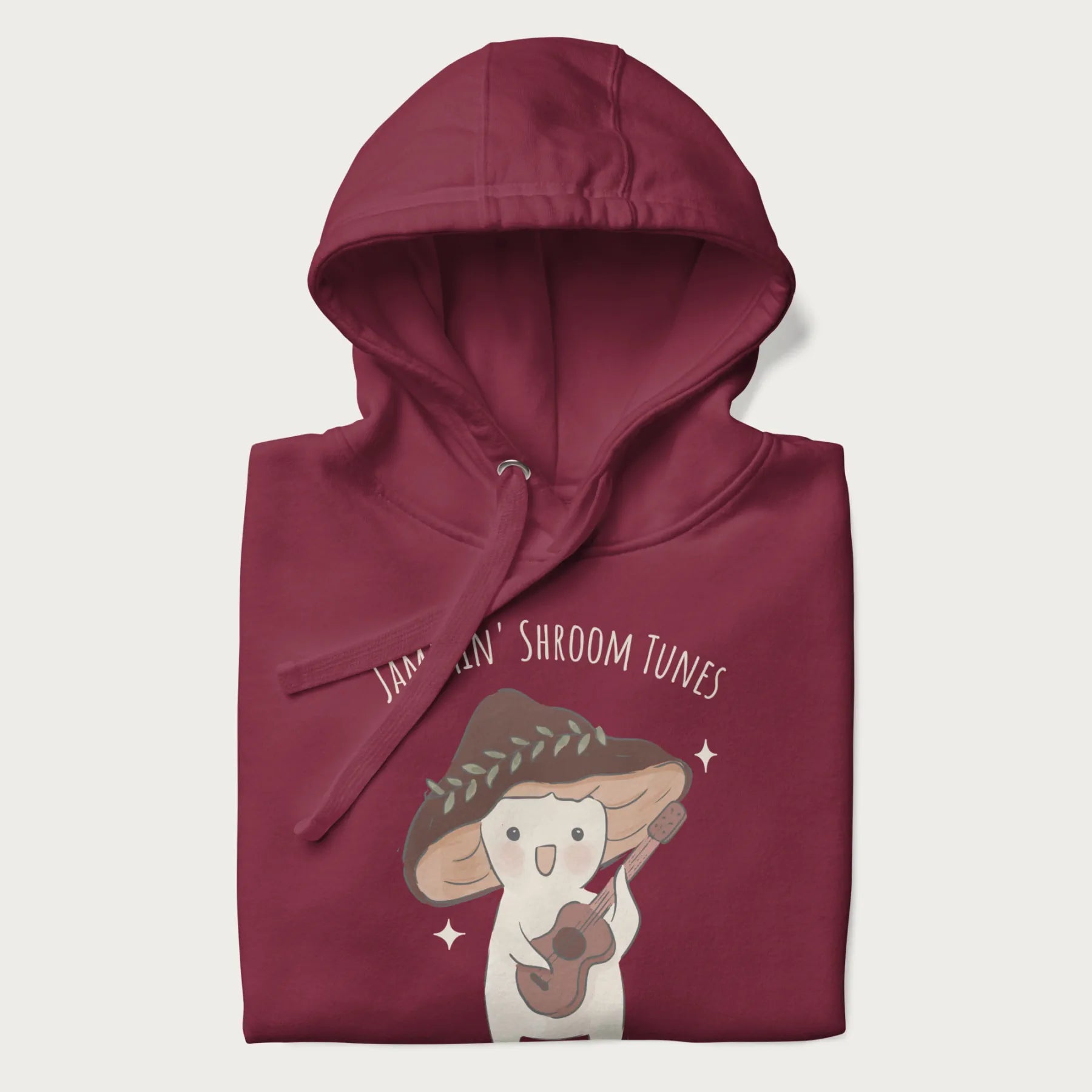 Folded maroon hoodie with a cute cottagecore mushroom character playing a guitar and the pun 'Jammin' Shroom Tunes' above.