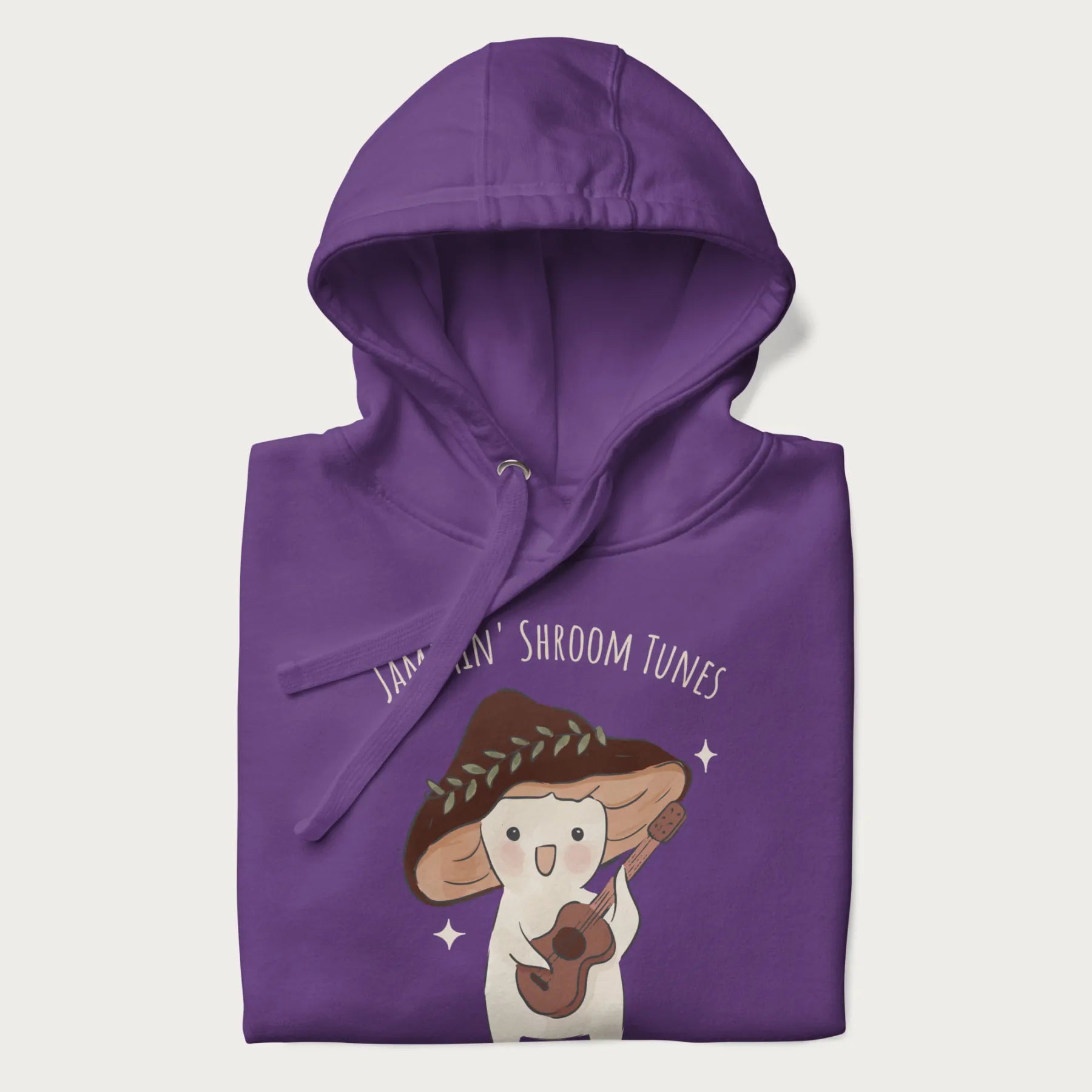 Folded purple hoodie with a cute cottagecore mushroom character playing a guitar and the pun 'Jammin' Shroom Tunes' above.