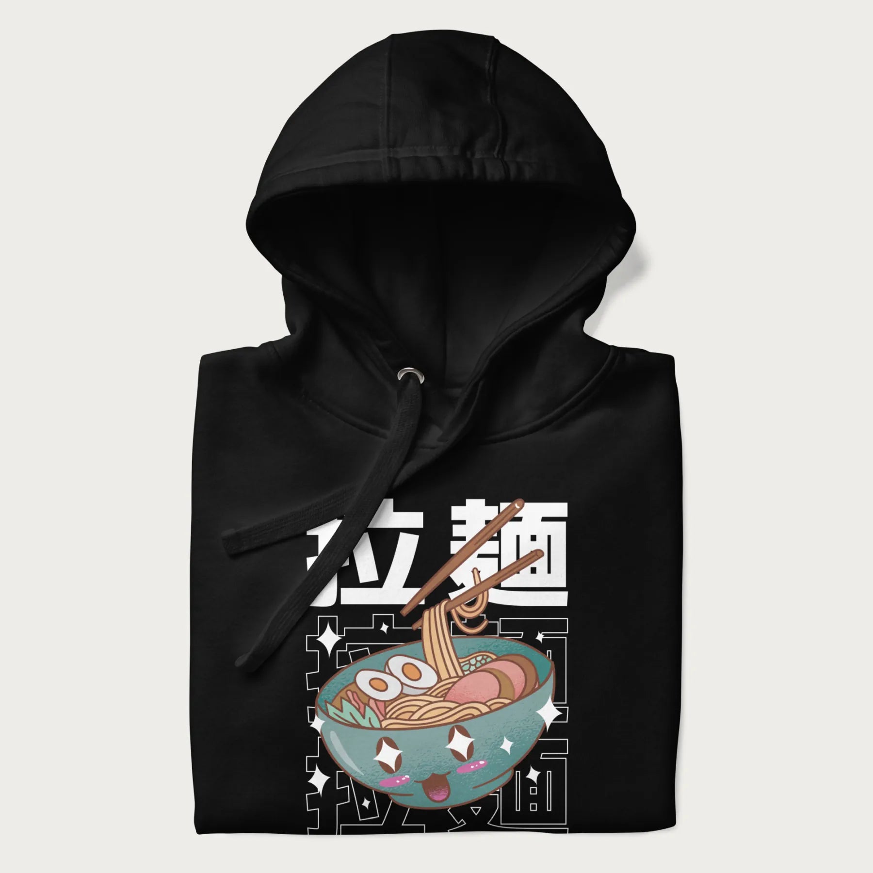 Folded black hoodie with Japanese text '拉麵' (Ramen) and graphic of a kawaii ramen bowl with sparkling eyes and blushed cheeks.