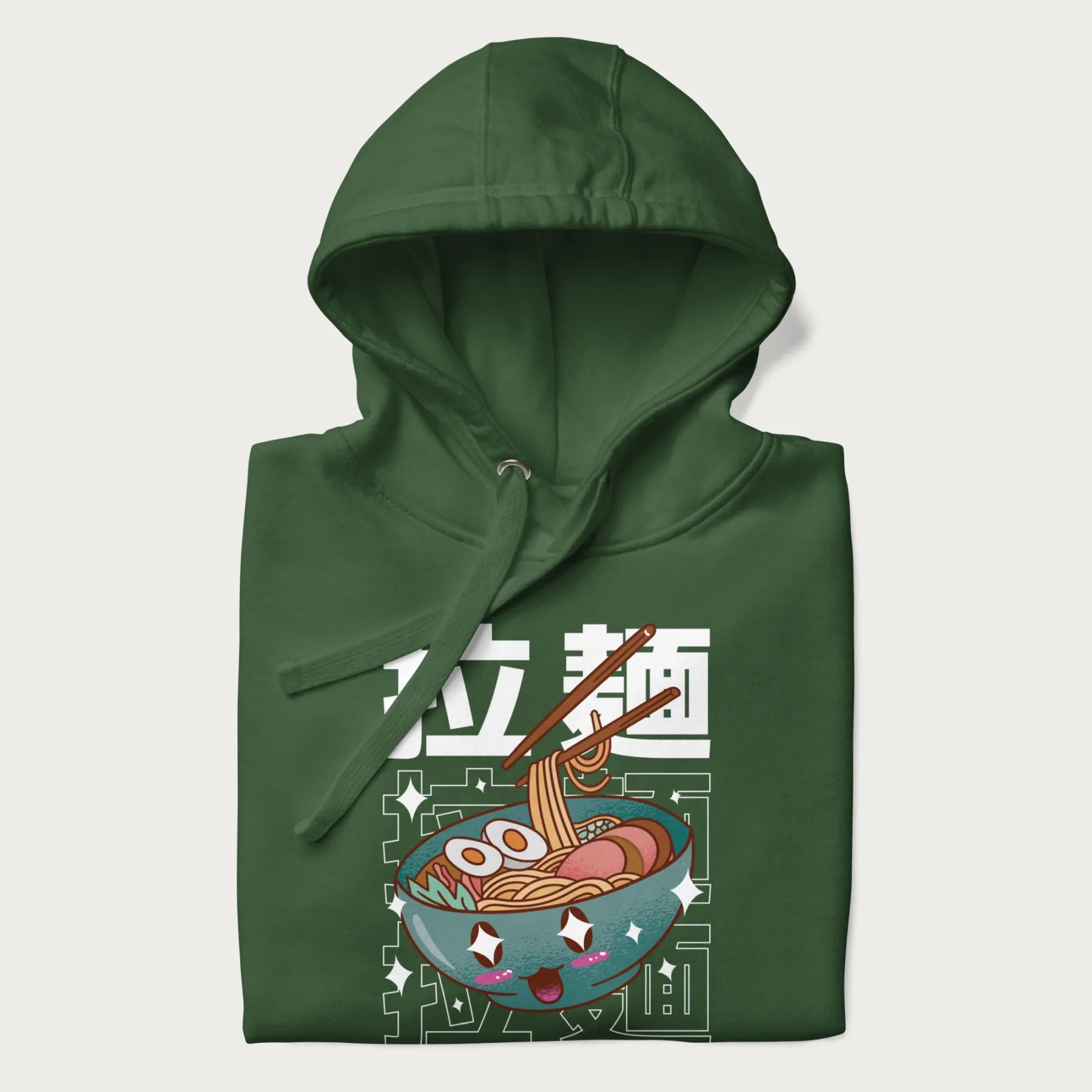 Folded forest green hoodie with Japanese text '拉麵' (Ramen) and graphic of a kawaii ramen bowl with sparkling eyes and blushed cheeks.
