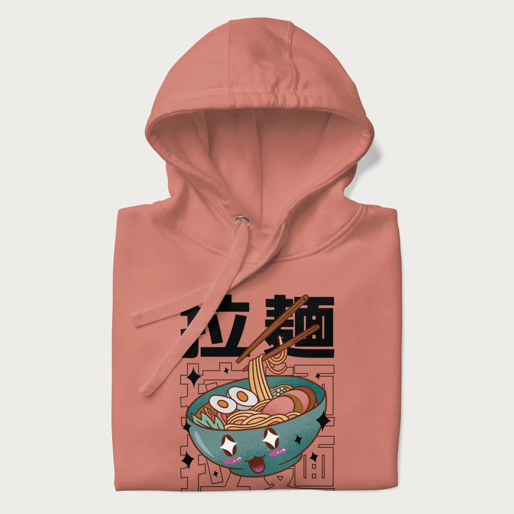 Folded light pink hoodie with Japanese text '拉麵' (Ramen) and graphic of a kawaii ramen bowl with sparkling eyes and blushed cheeks.