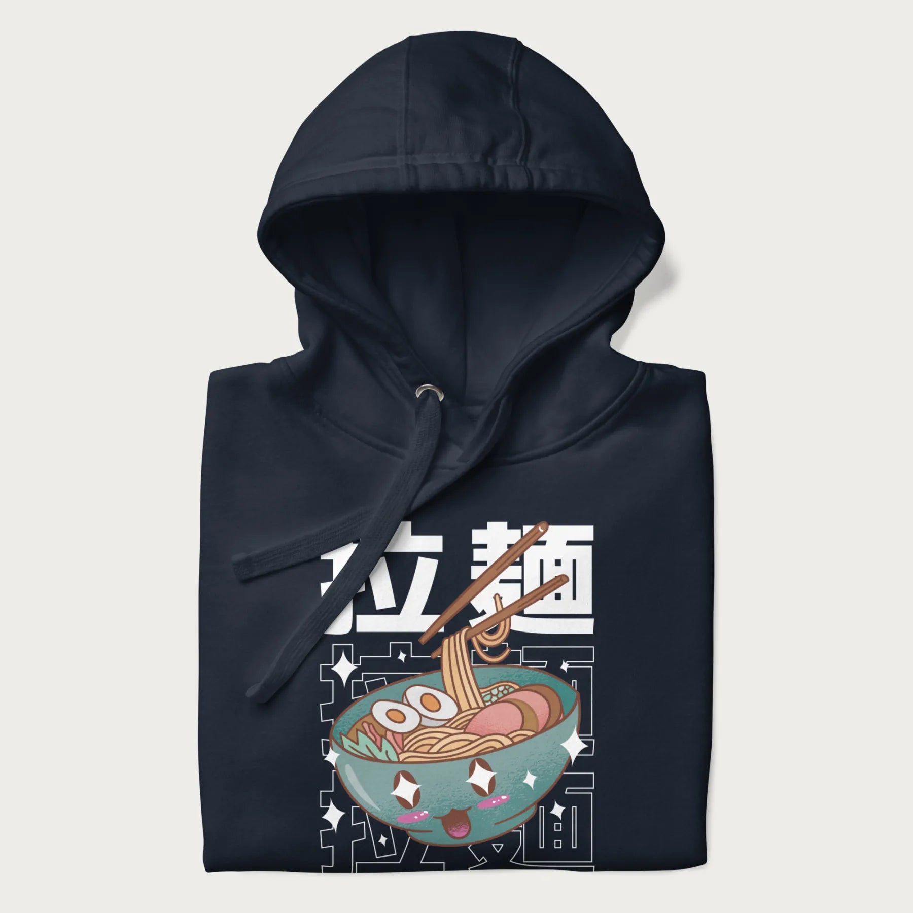 Folded navy blue hoodie with Japanese text '拉麵' (Ramen) and graphic of a kawaii ramen bowl with sparkling eyes and blushed cheeks.