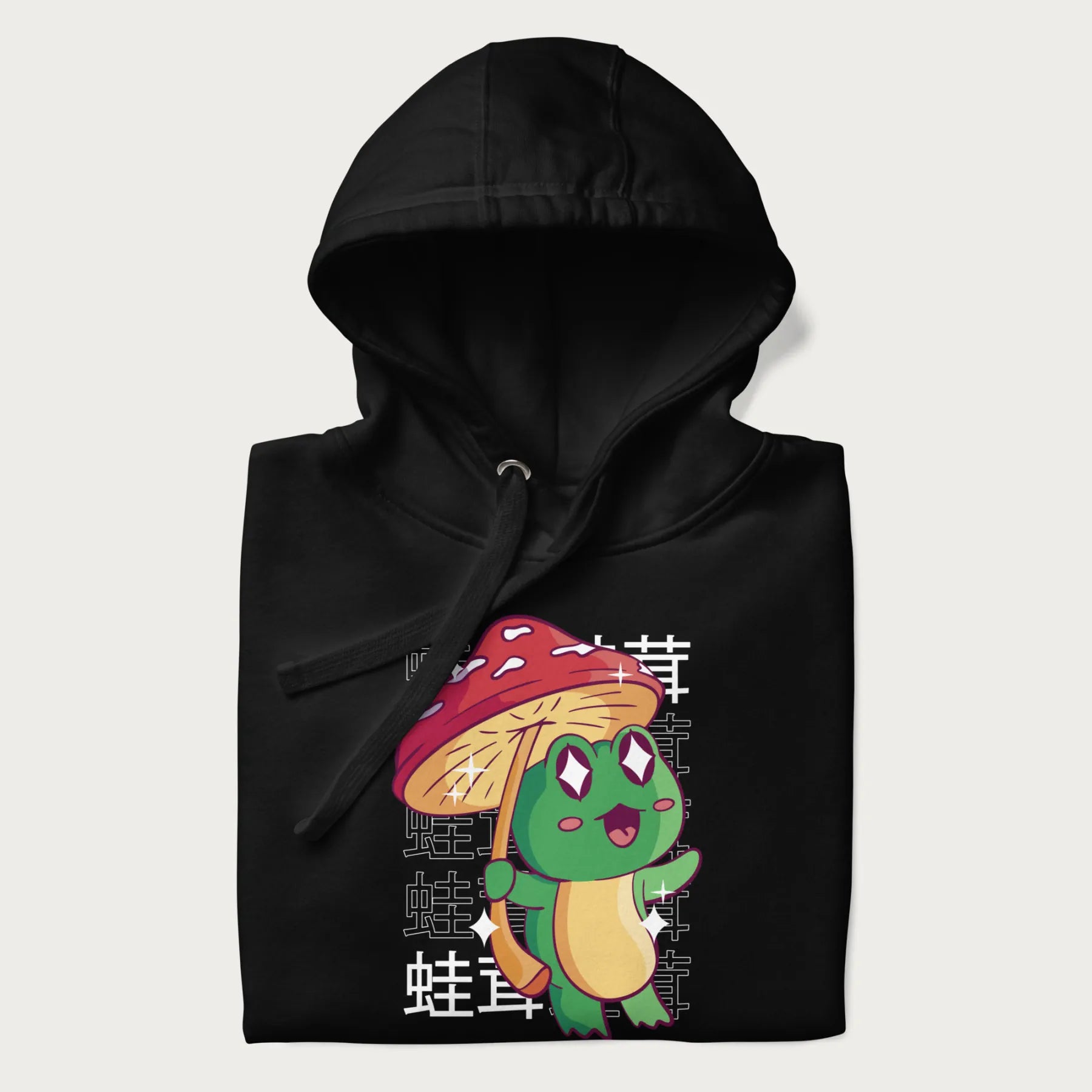 Folded black hoodie with Japanese graphic of a cute green frog holding a red mushroom and Japanese text '蛙茸' in the background.
