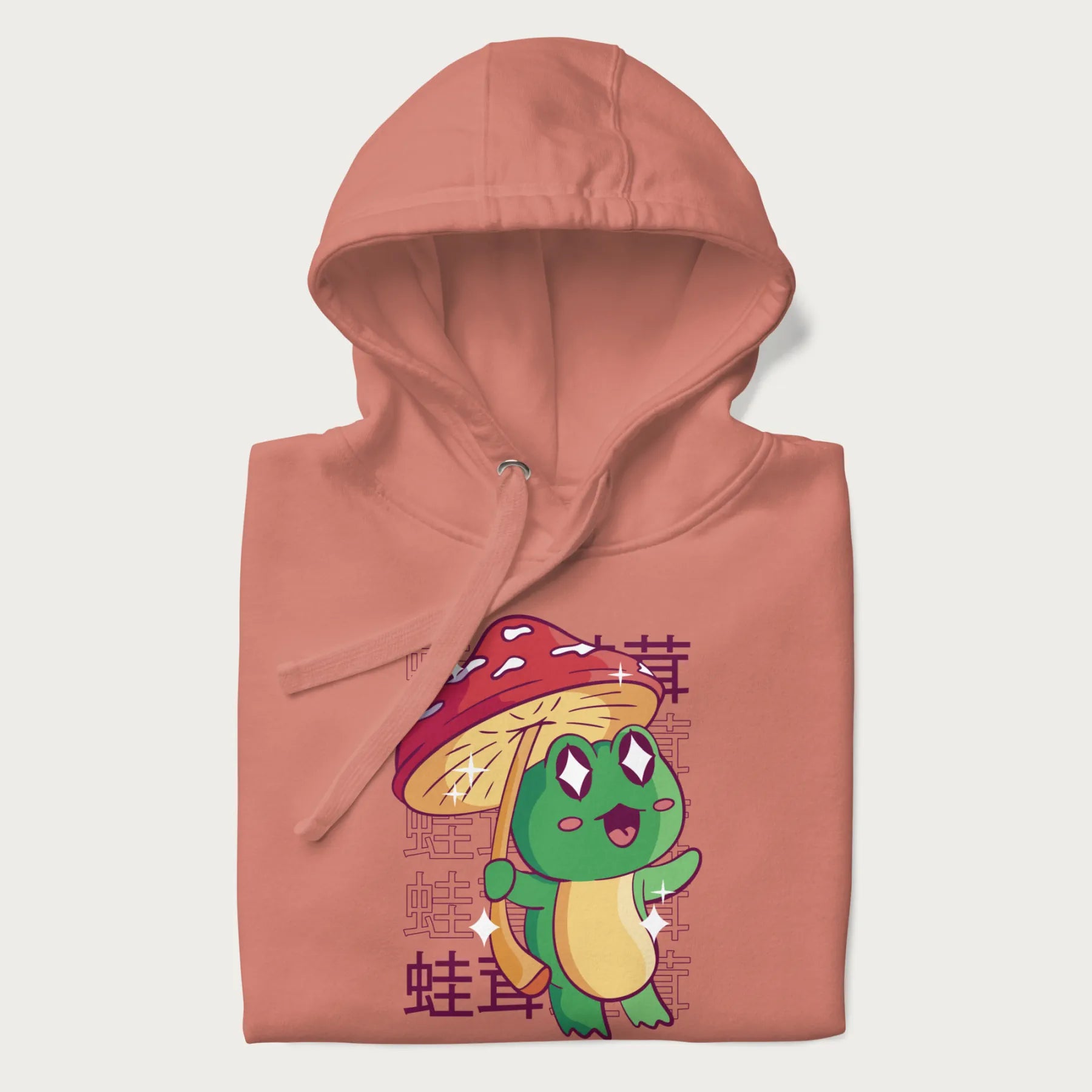 Folded light pink hoodie with Japanese graphic of a cute green frog holding a red mushroom and Japanese text '蛙茸' in the background.