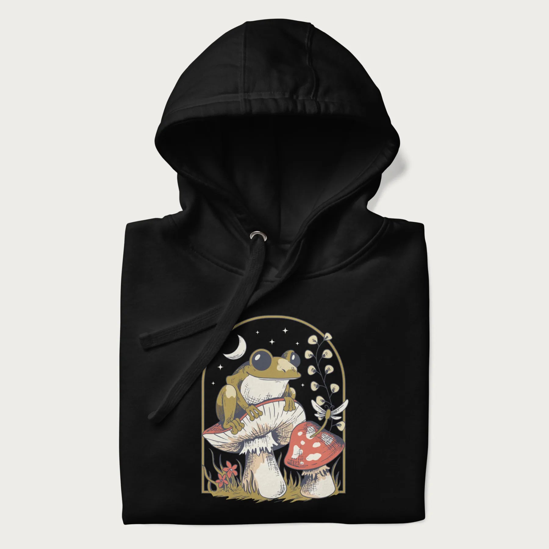 Folded black hoodie with cottagecore graphic of a frog perched on a mushroom, surrounded by additional mushrooms, flowers, and a crescent moon with stars.