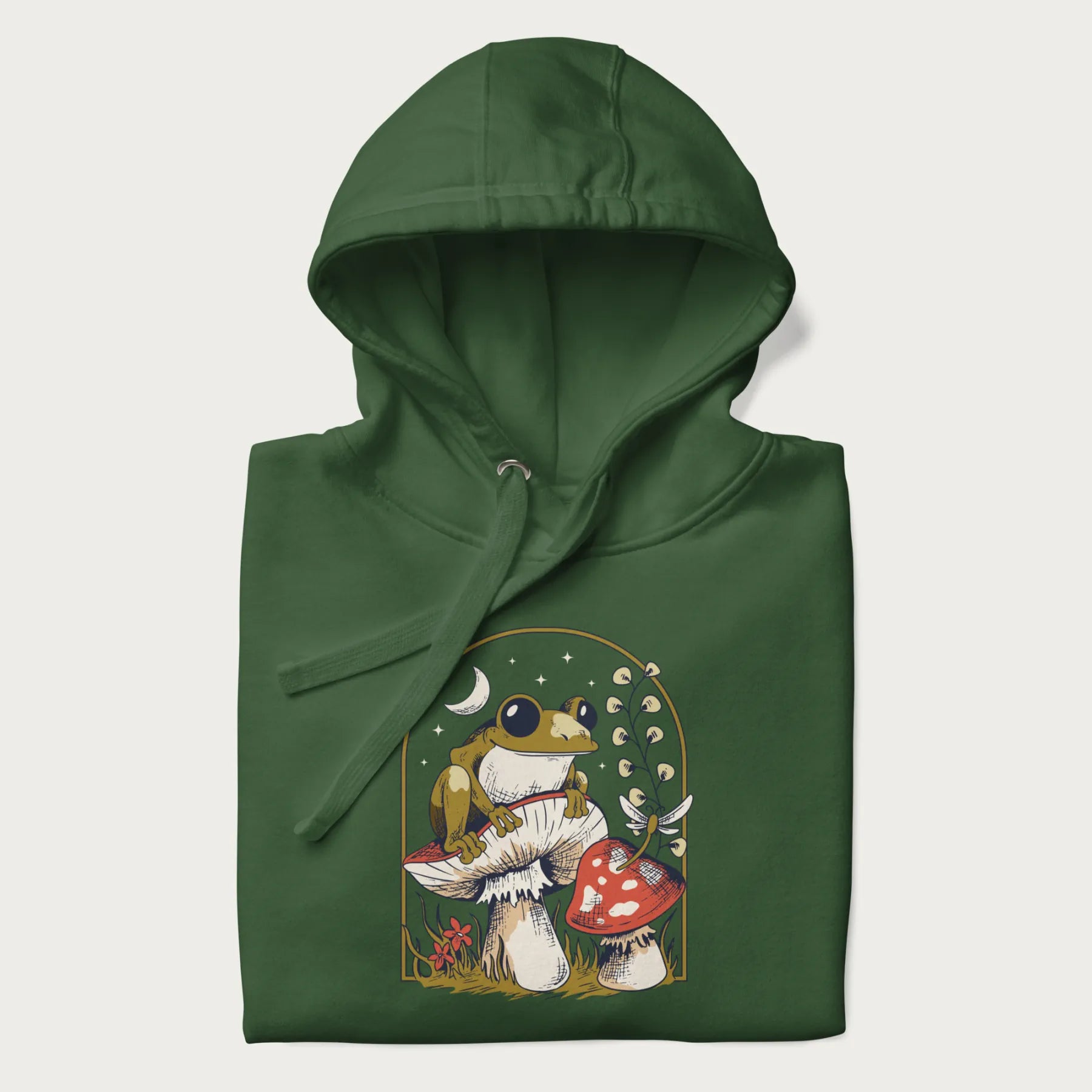 Folded forest green hoodie with cottagecore graphic of a frog perched on a mushroom, surrounded by additional mushrooms, flowers, and a crescent moon with stars.