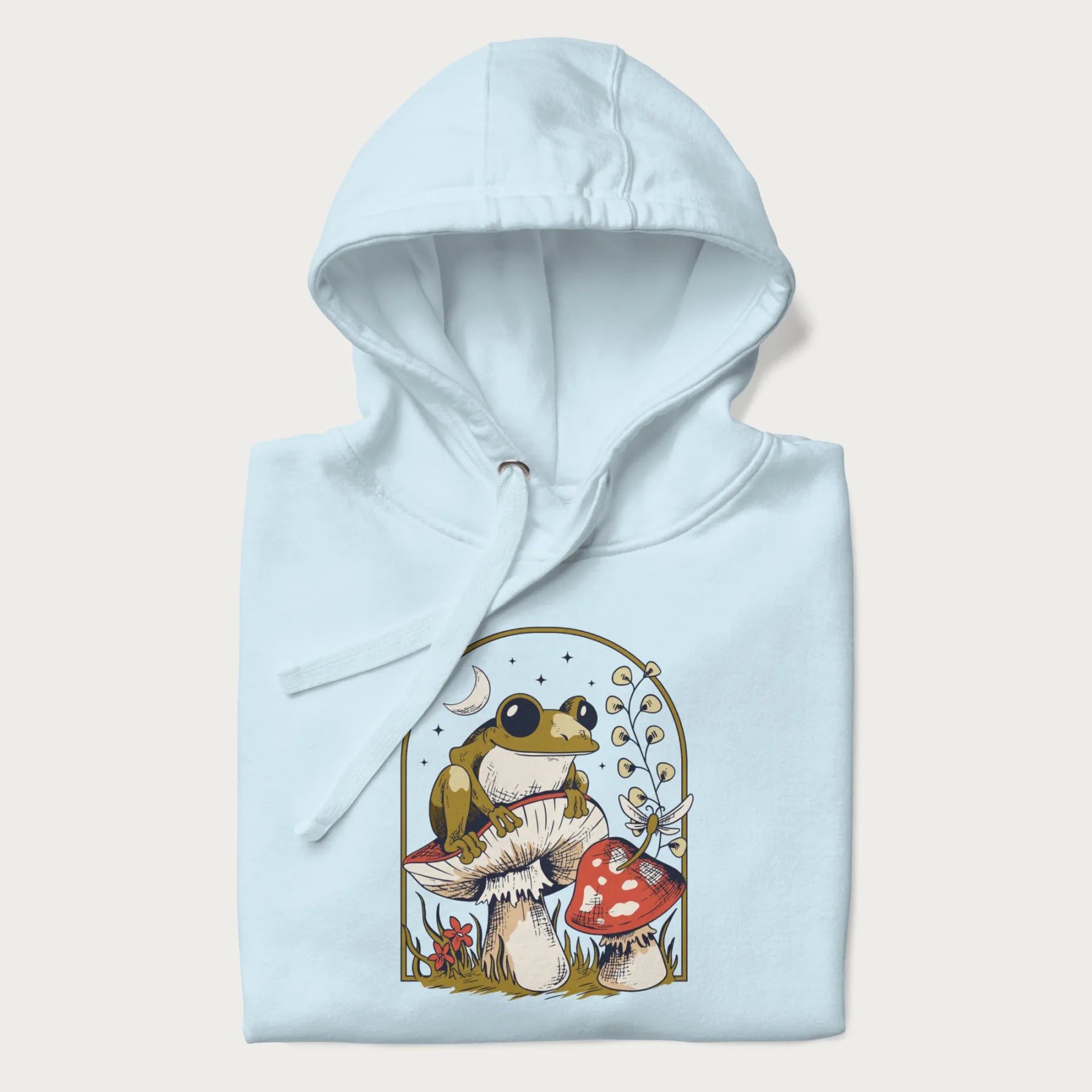 Folded light blue hoodie with cottagecore graphic of a frog perched on a mushroom, surrounded by additional mushrooms, flowers, and a crescent moon with stars.