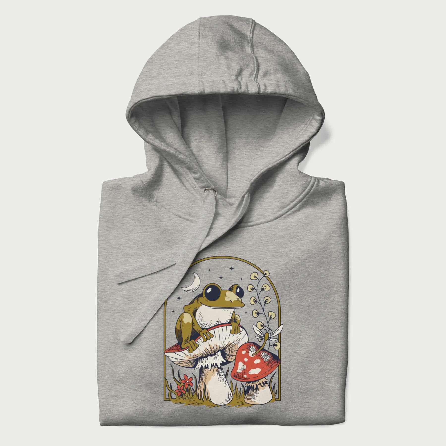 Folded light grey hoodie with cottagecore graphic of a frog perched on a mushroom, surrounded by additional mushrooms, flowers, and a crescent moon with stars.