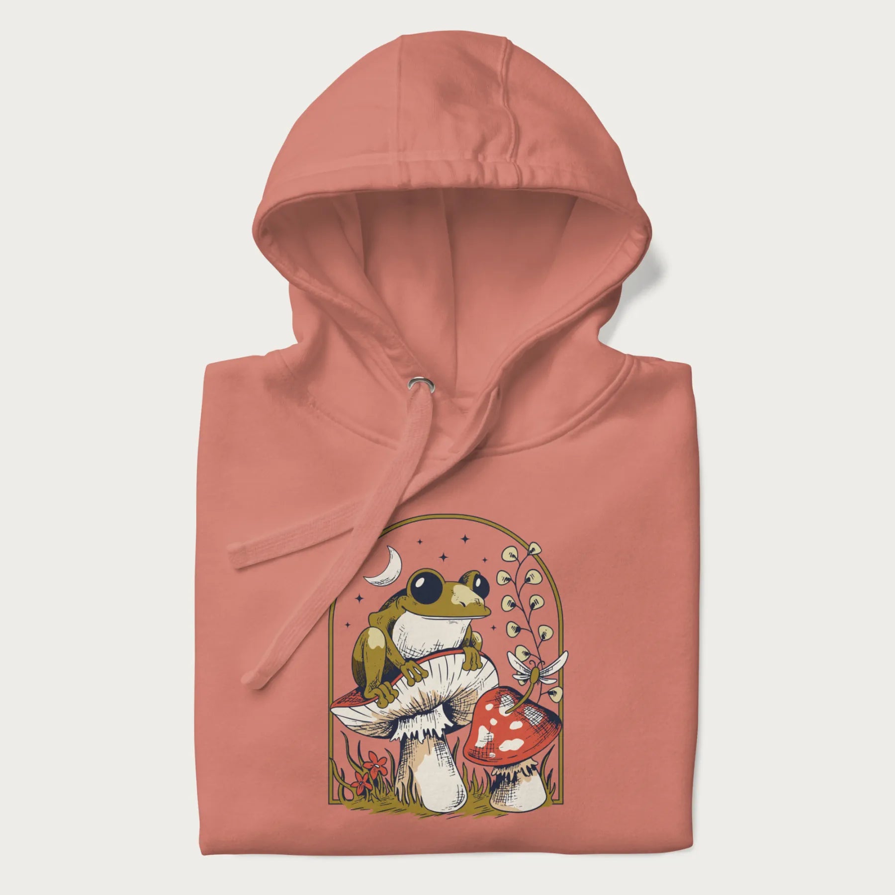 Folded light pink hoodie with cottagecore graphic of a frog perched on a mushroom, surrounded by additional mushrooms, flowers, and a crescent moon with stars.