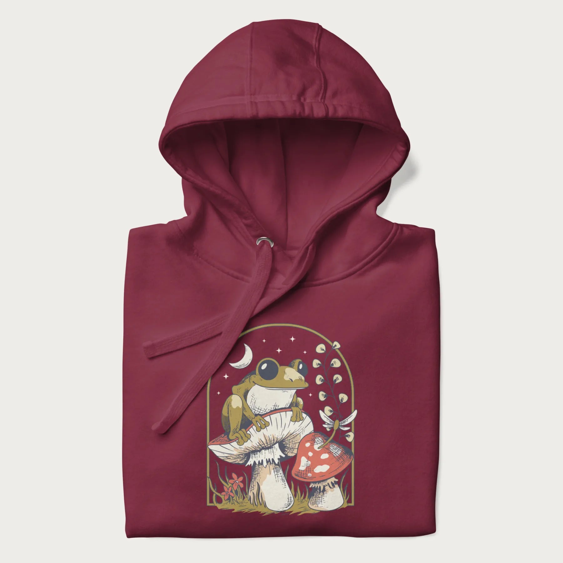 Folded maroon hoodie with cottagecore graphic of a frog perched on a mushroom, surrounded by additional mushrooms, flowers, and a crescent moon with stars.