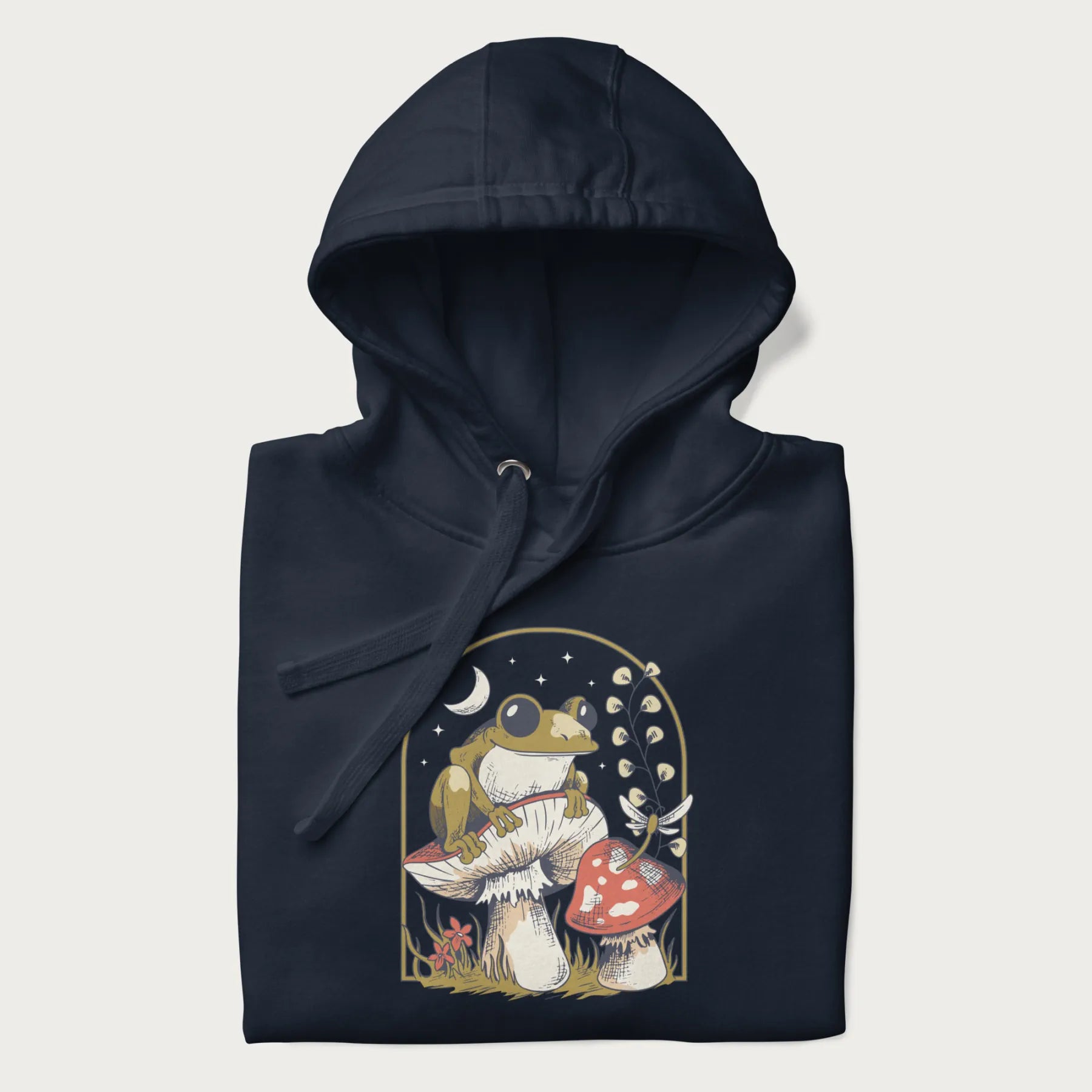 Folded navy blue hoodie with cottagecore graphic of a frog perched on a mushroom, surrounded by additional mushrooms, flowers, and a crescent moon with stars.
