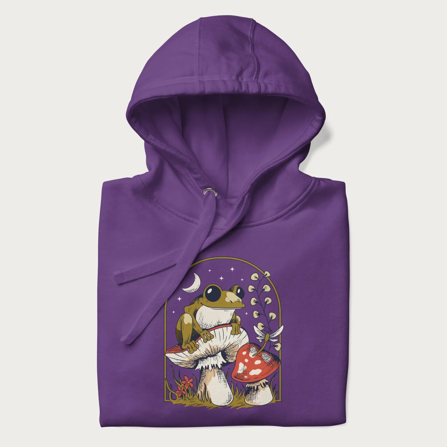 Folded purple hoodie with cottagecore graphic of a frog perched on a mushroom, surrounded by additional mushrooms, flowers, and a crescent moon with stars.