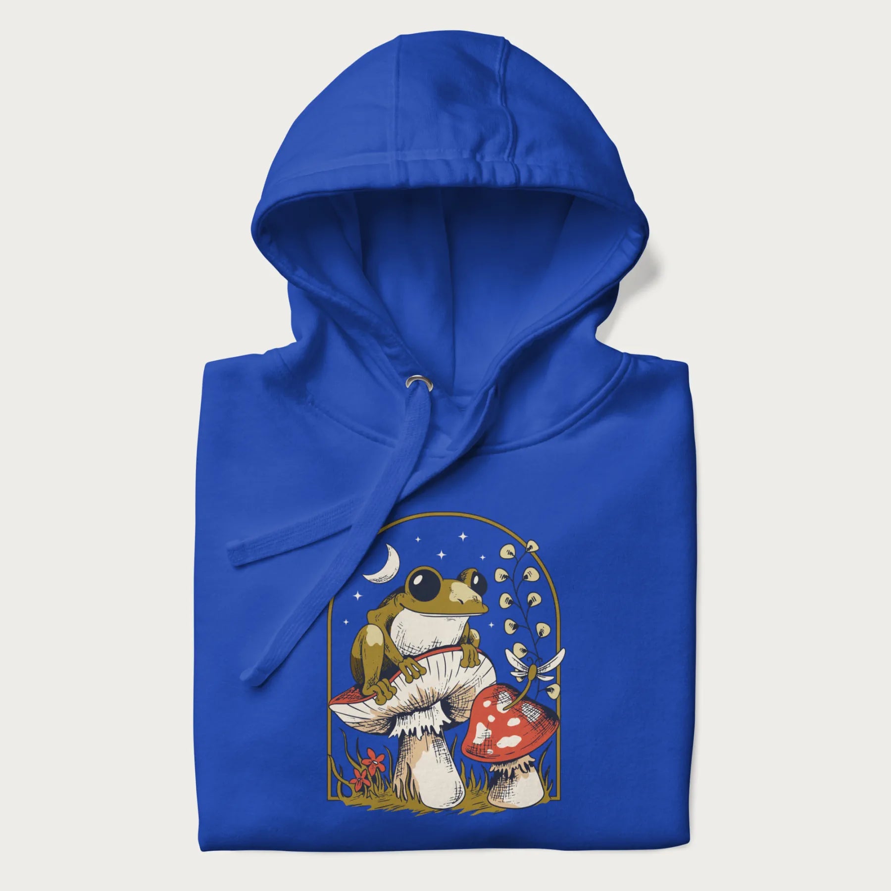 Folded royal blue hoodie with cottagecore graphic of a frog perched on a mushroom, surrounded by additional mushrooms, flowers, and a crescent moon with stars.