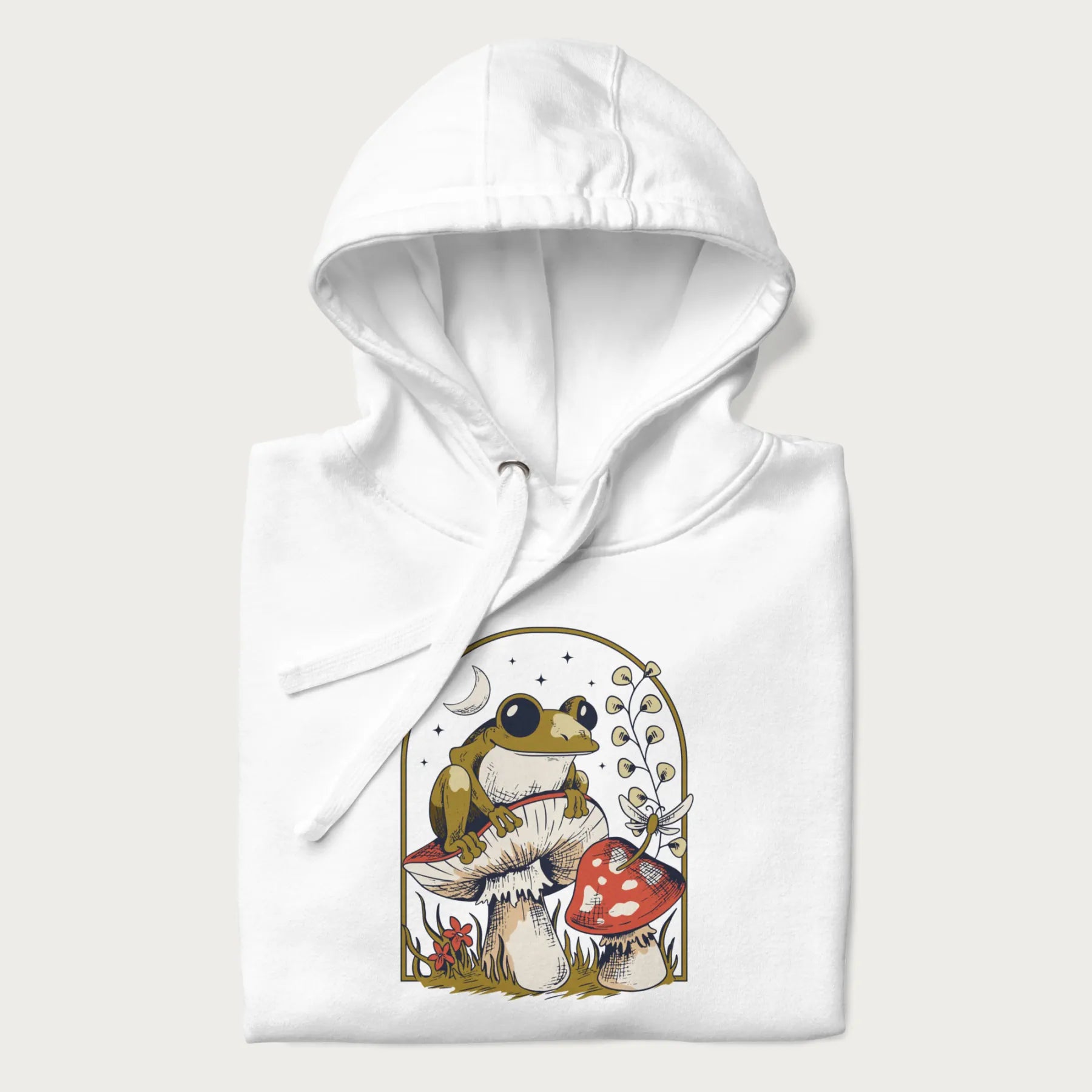 Folded white hoodie with cottagecore graphic of a frog perched on a mushroom, surrounded by additional mushrooms, flowers, and a crescent moon with stars.
