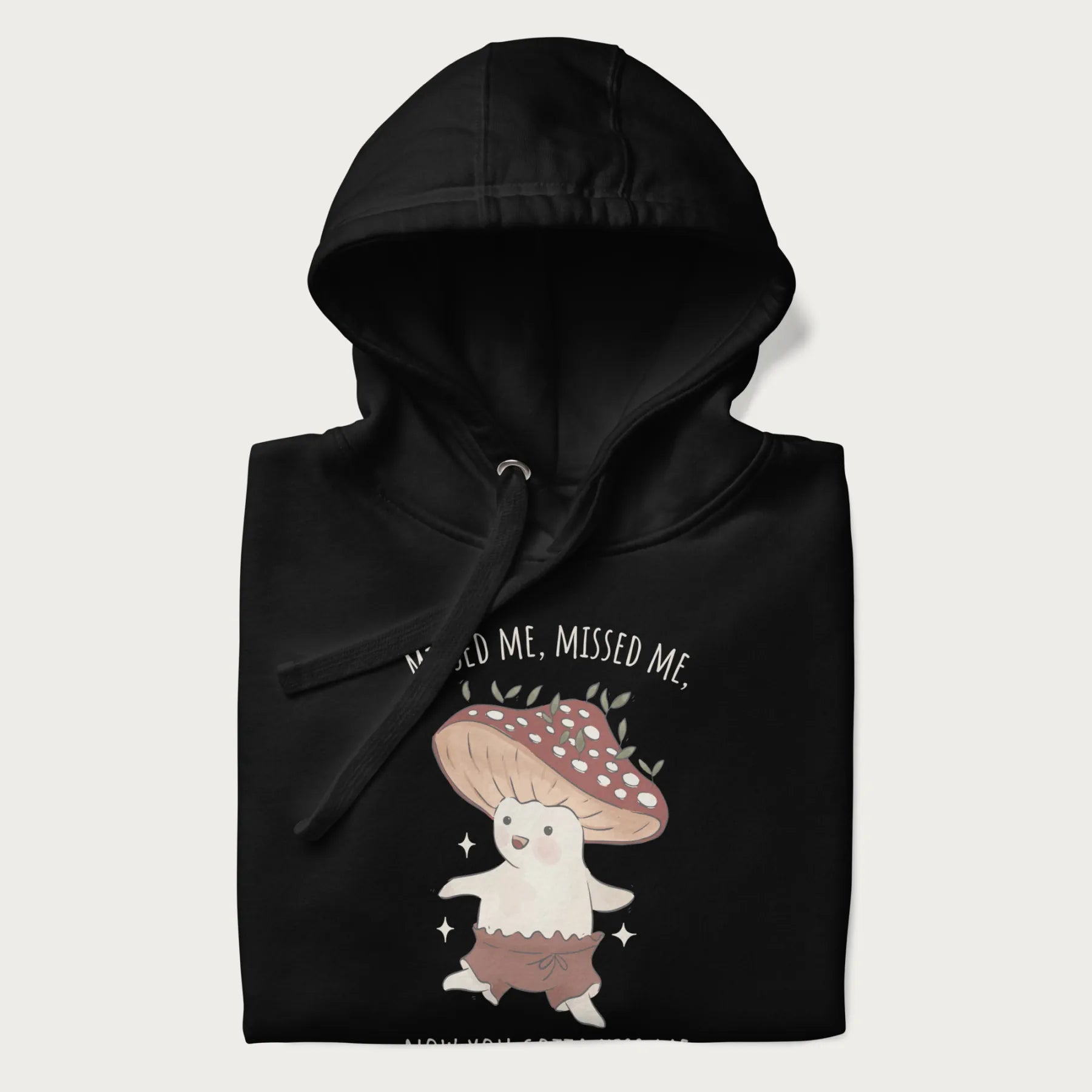 Folded black hoodie with a graphic of a cute cottagecore mushroom character dancing and the phrase 'Missed Me, Missed Me, Now You Gotta Kiss Me'.