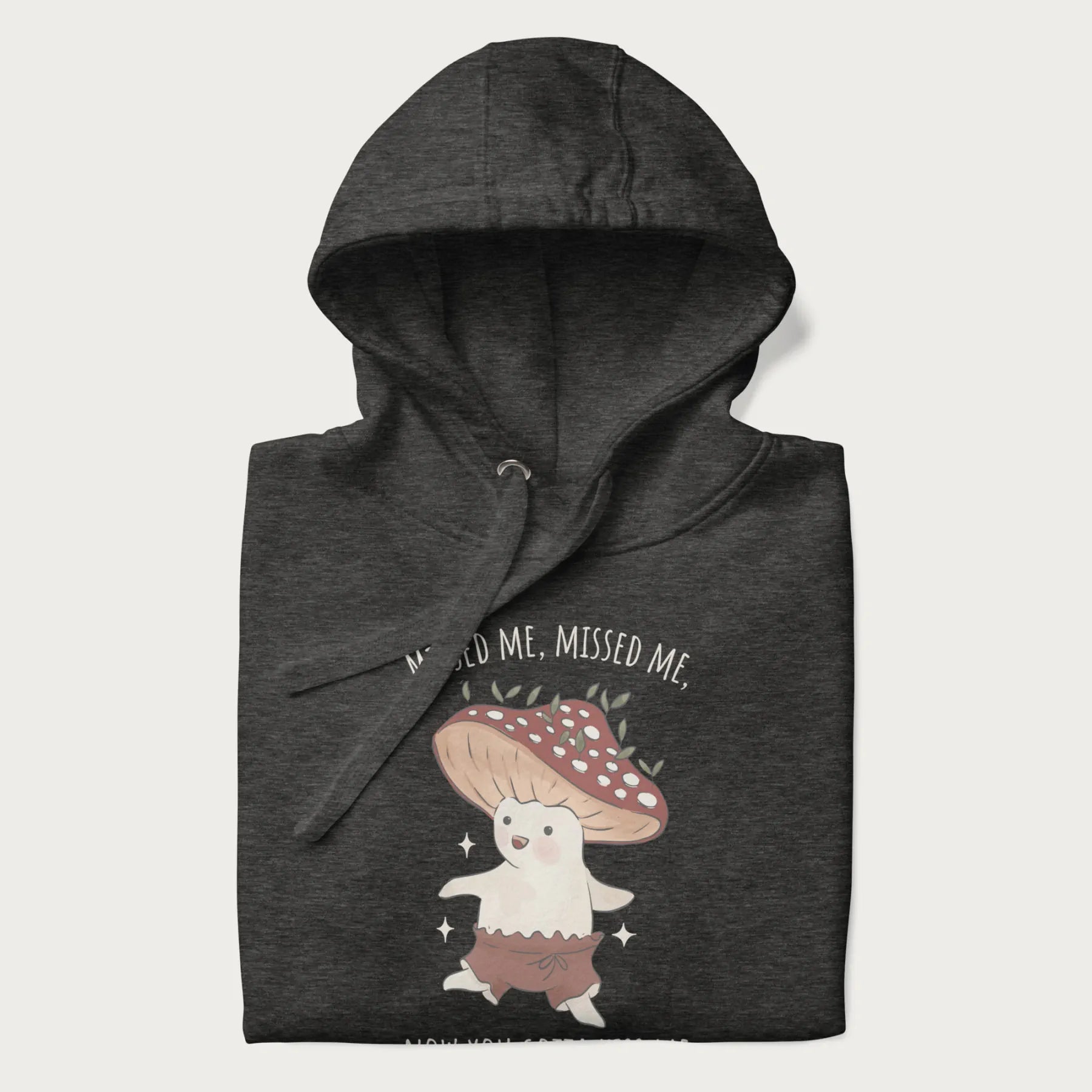 Folded dark grey hoodie with a graphic of a cute cottagecore mushroom character dancing and the phrase 'Missed Me, Missed Me, Now You Gotta Kiss Me'.