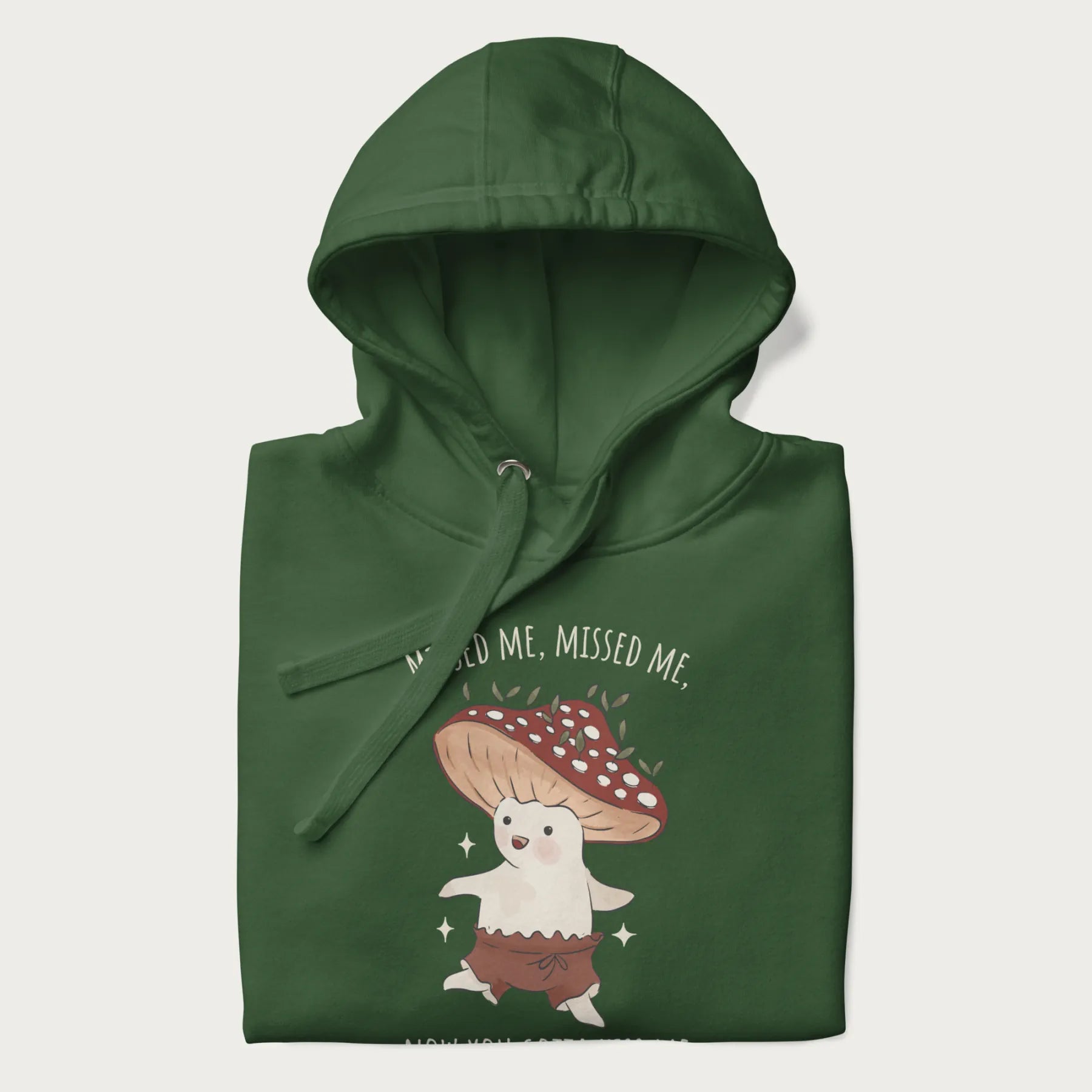 Folded forest green hoodie with a graphic of a cute cottagecore mushroom character dancing and the phrase 'Missed Me, Missed Me, Now You Gotta Kiss Me'.