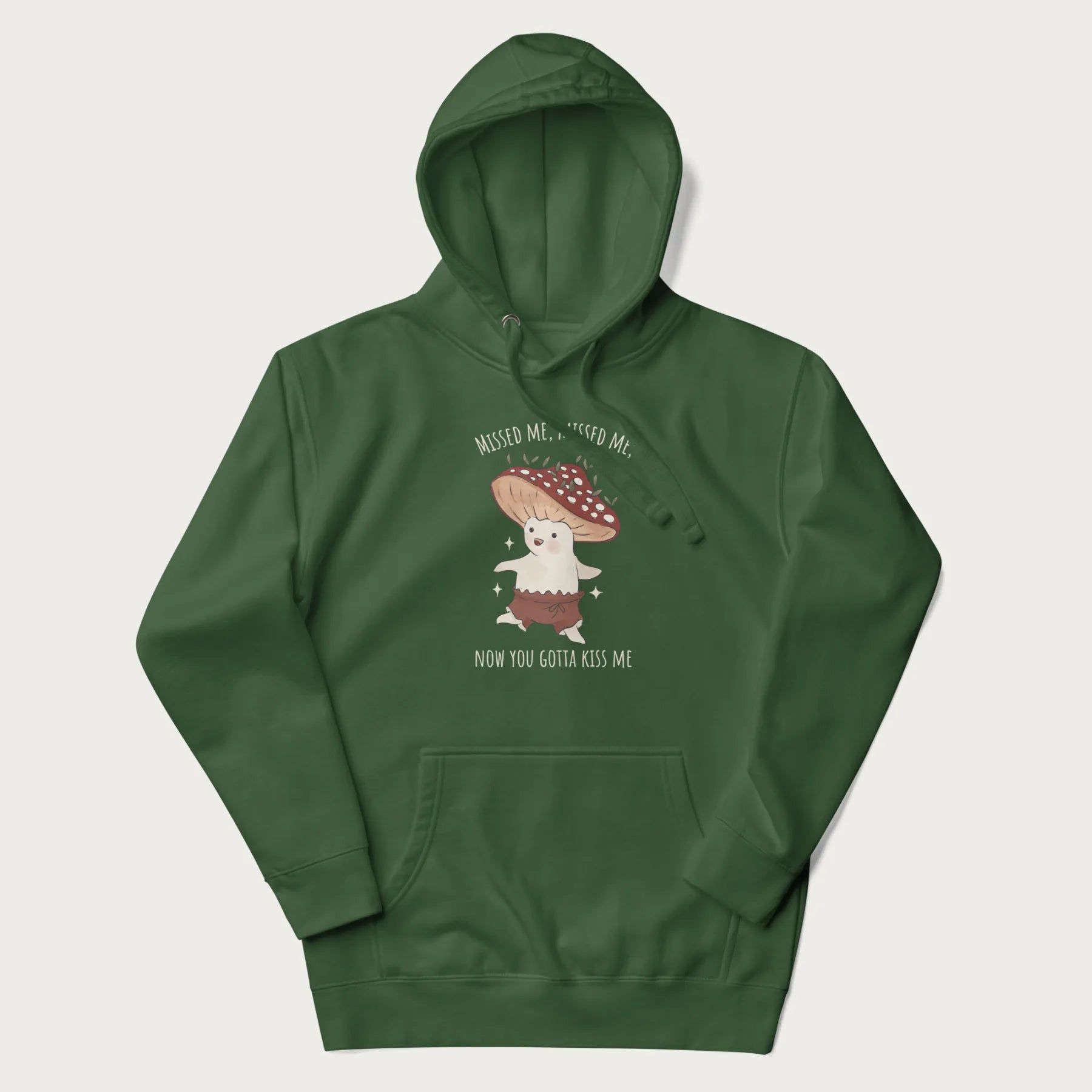 Forest green hoodie with a graphic of a cute cottagecore mushroom character dancing and the phrase 'Missed Me, Missed Me, Now You Gotta Kiss Me'.