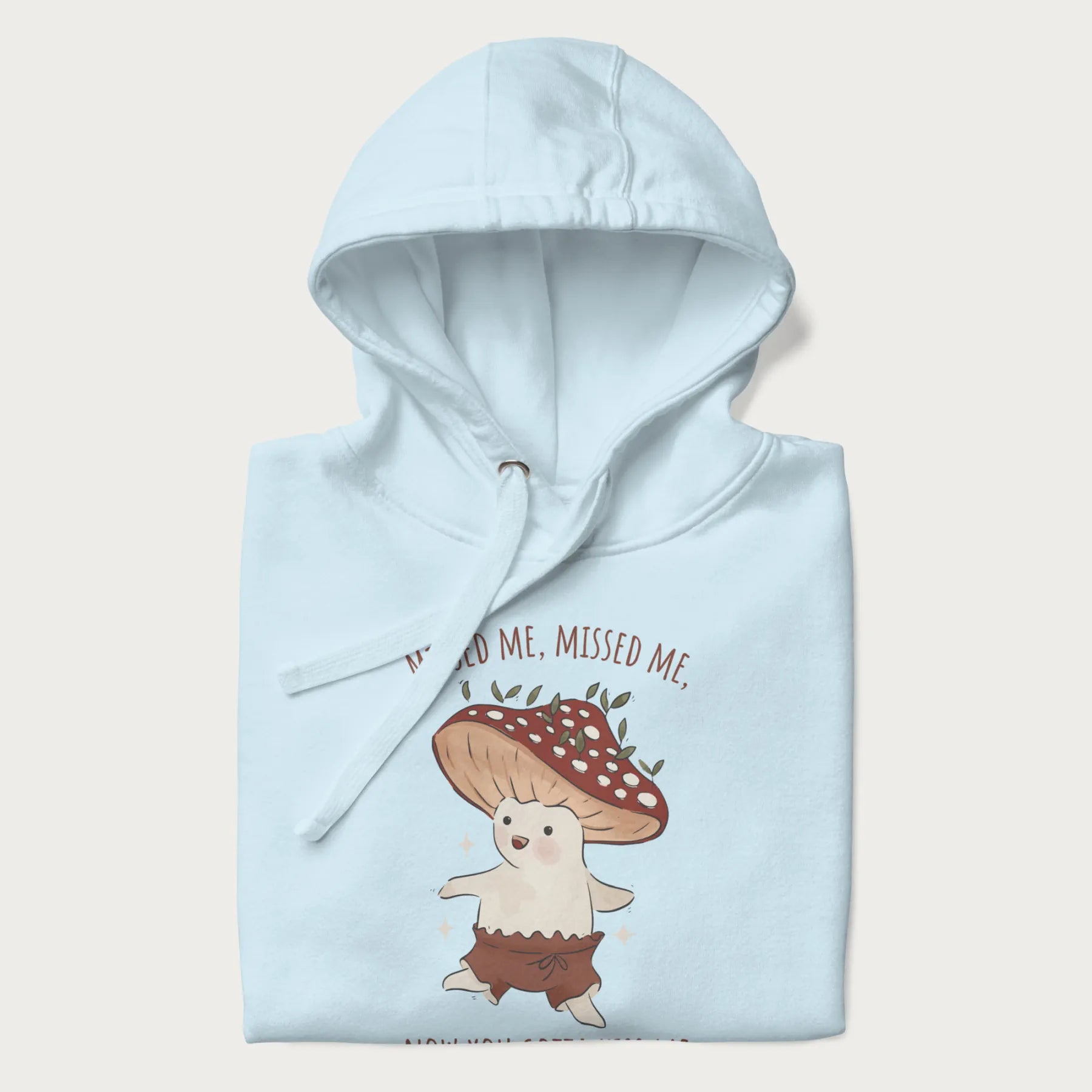 Folded light blue hoodie with a graphic of a cute cottagecore mushroom character dancing and the phrase 'Missed Me, Missed Me, Now You Gotta Kiss Me'.