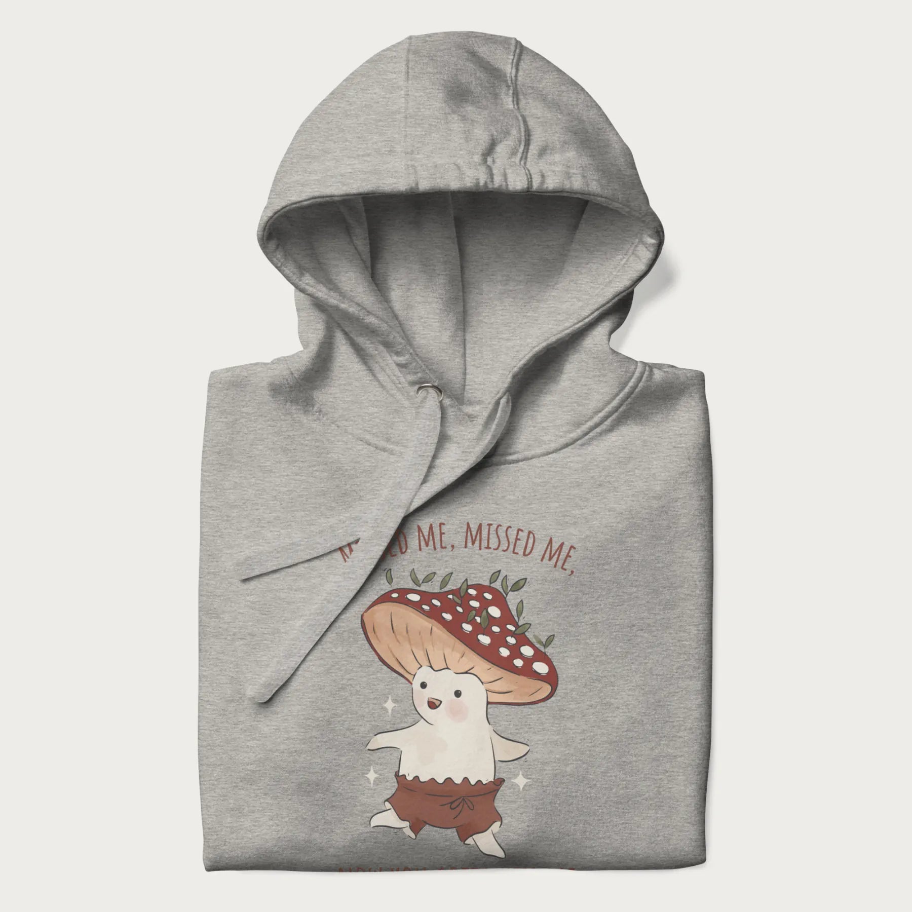 Folded light grey hoodie with a graphic of a cute cottagecore mushroom character dancing and the phrase 'Missed Me, Missed Me, Now You Gotta Kiss Me'.