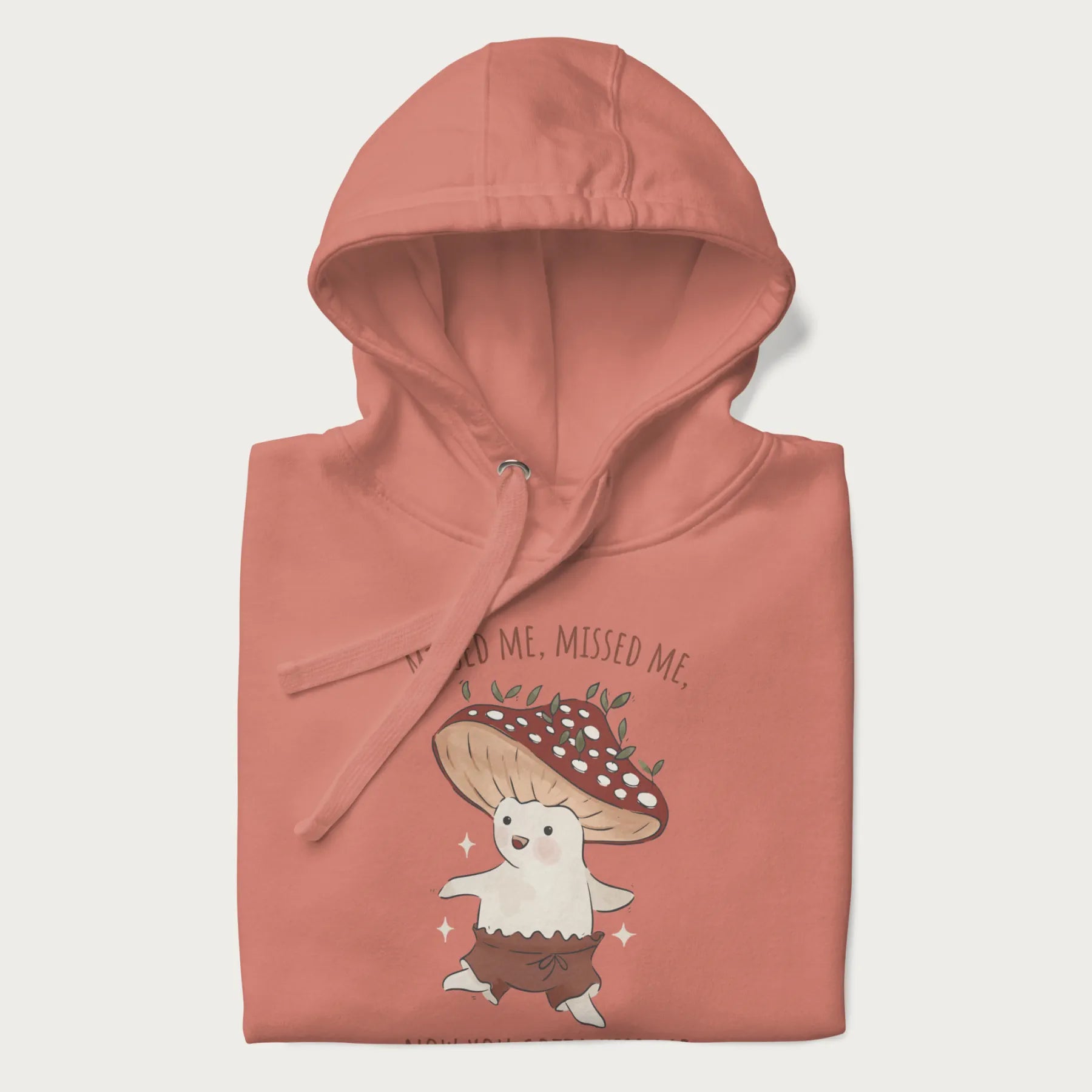 Folded light pink hoodie with a graphic of a cute cottagecore mushroom character dancing and the phrase 'Missed Me, Missed Me, Now You Gotta Kiss Me'.
