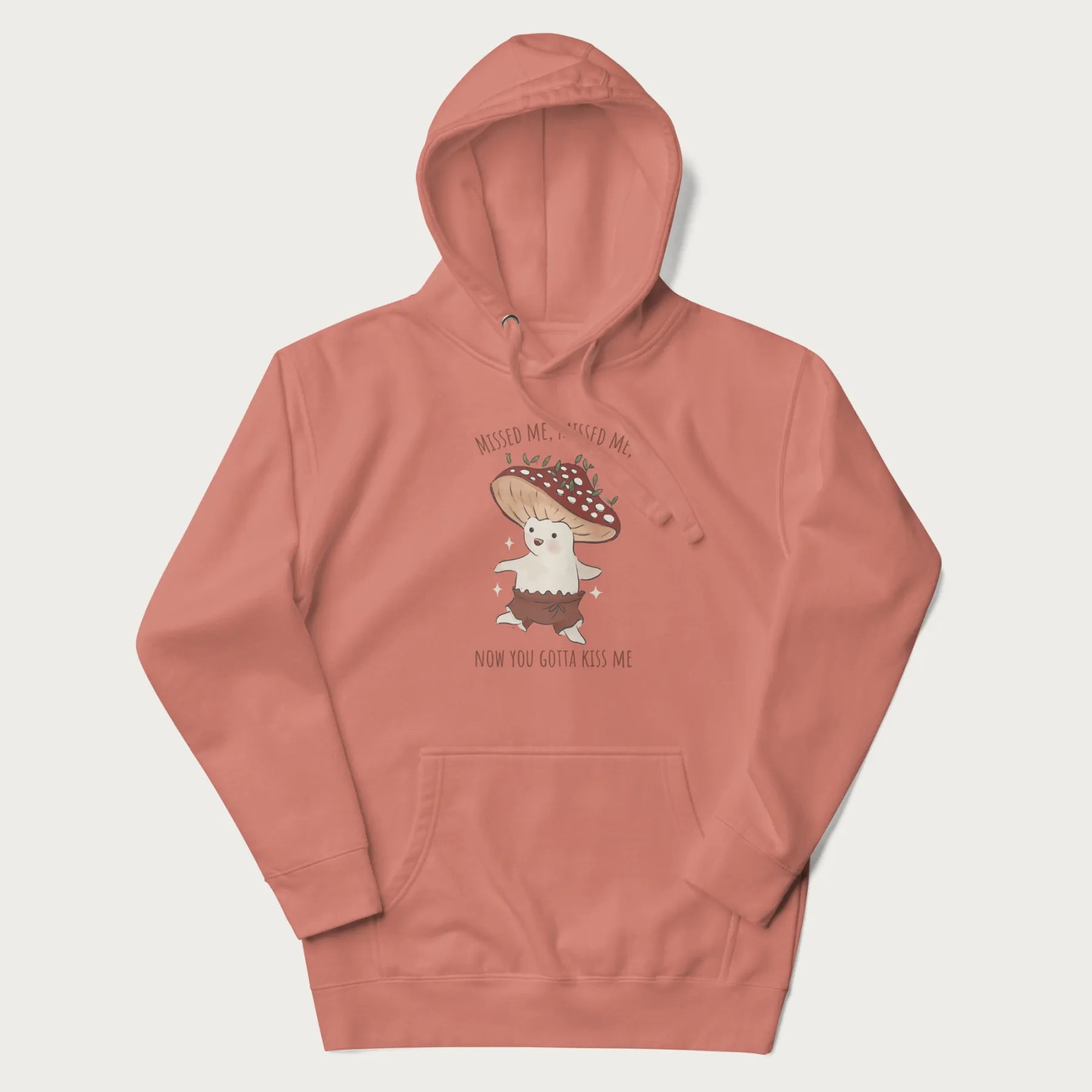 Light pink hoodie with a graphic of a cute cottagecore mushroom character dancing and the phrase 'Missed Me, Missed Me, Now You Gotta Kiss Me'.
