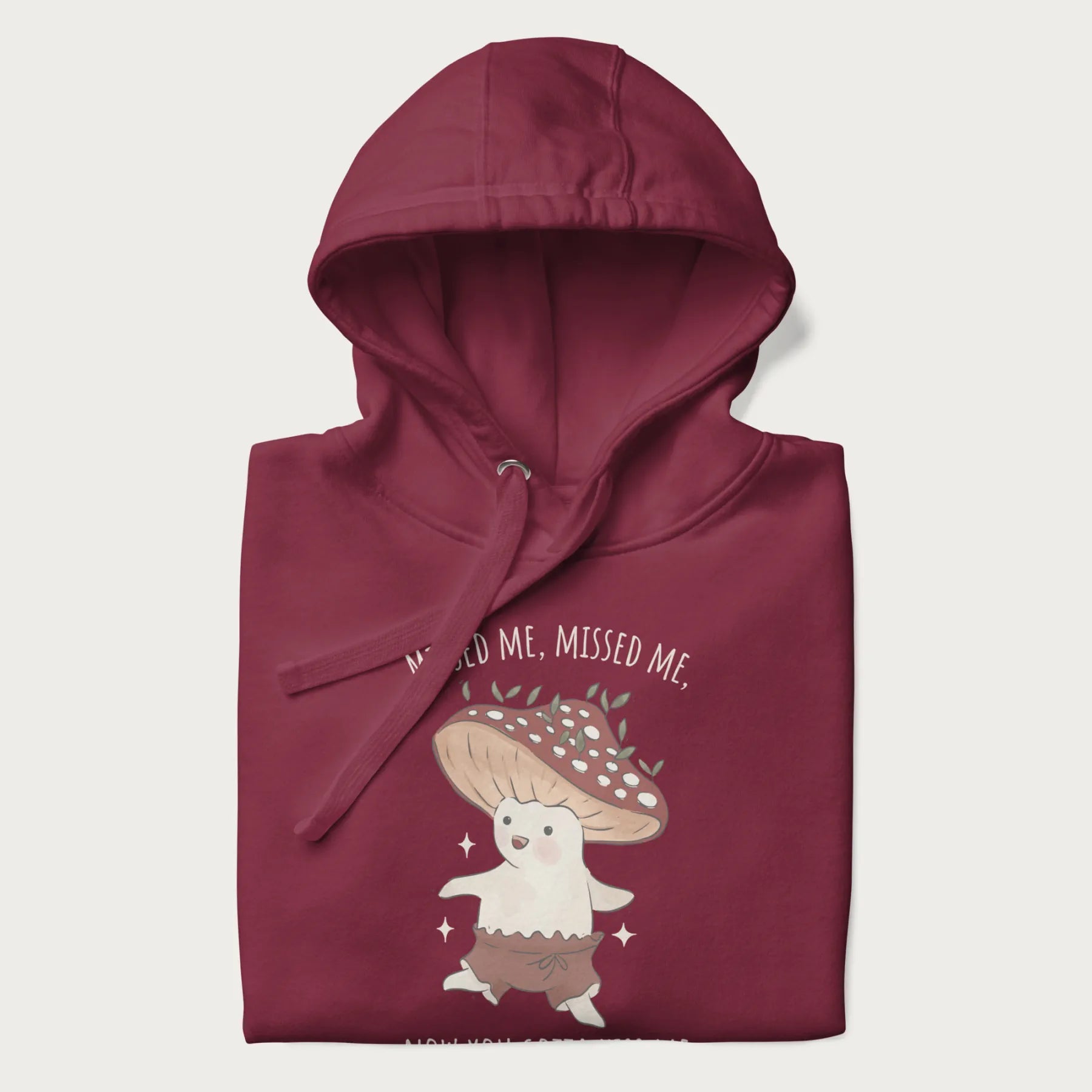 Folded maroon hoodie with a graphic of a cute cottagecore mushroom character dancing and the phrase 'Missed Me, Missed Me, Now You Gotta Kiss Me'.