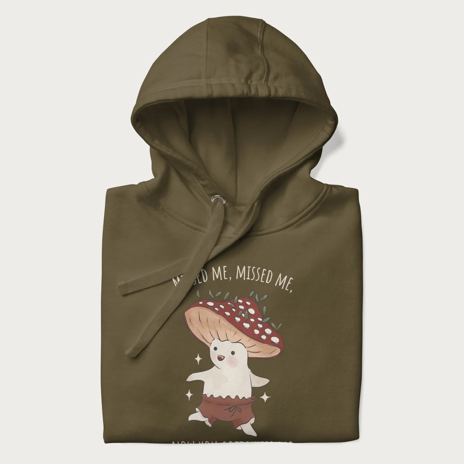 Folded military green hoodie with a graphic of a cute cottagecore mushroom character dancing and the phrase 'Missed Me, Missed Me, Now You Gotta Kiss Me'.