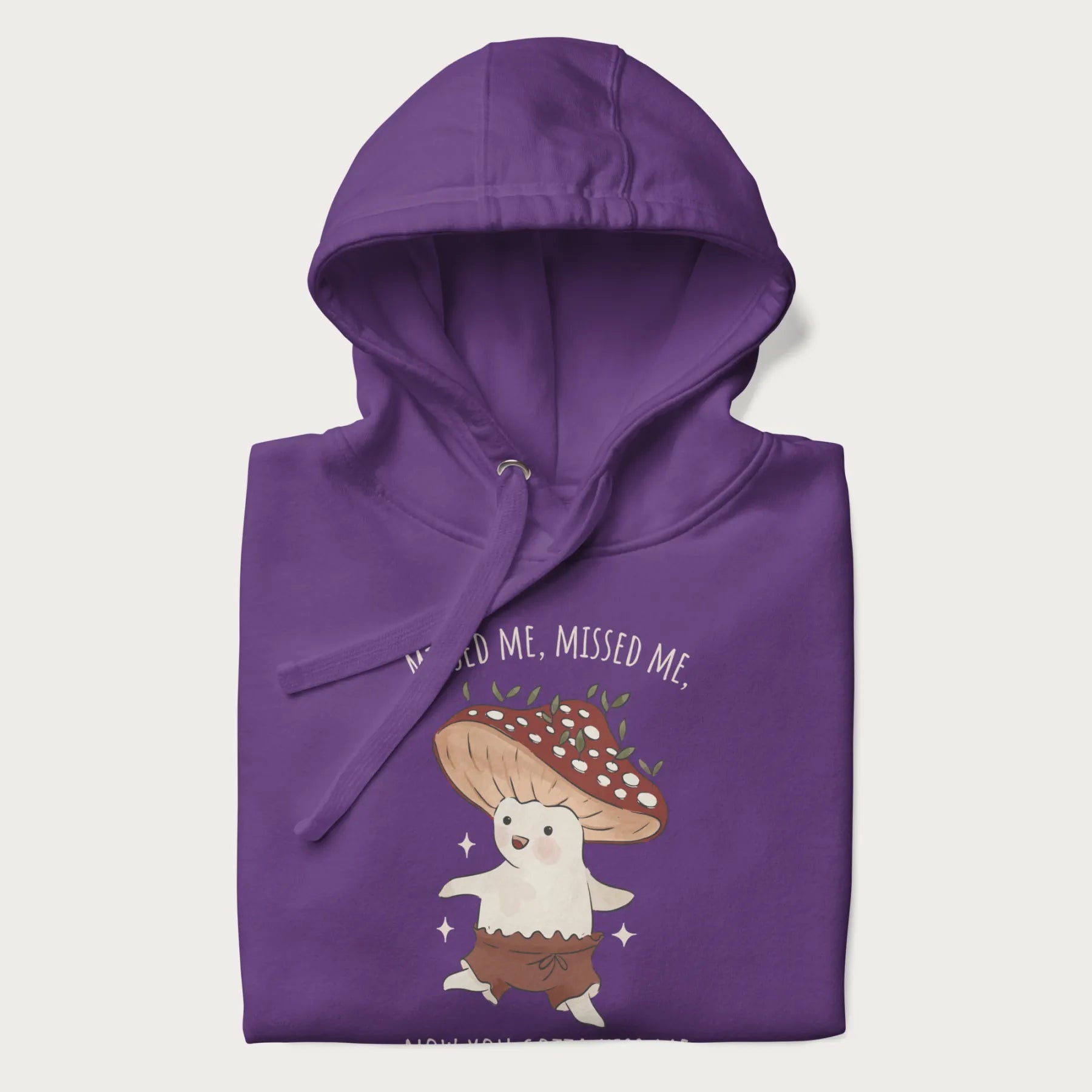 Folded purple hoodie with a graphic of a cute cottagecore mushroom character dancing and the phrase 'Missed Me, Missed Me, Now You Gotta Kiss Me'.