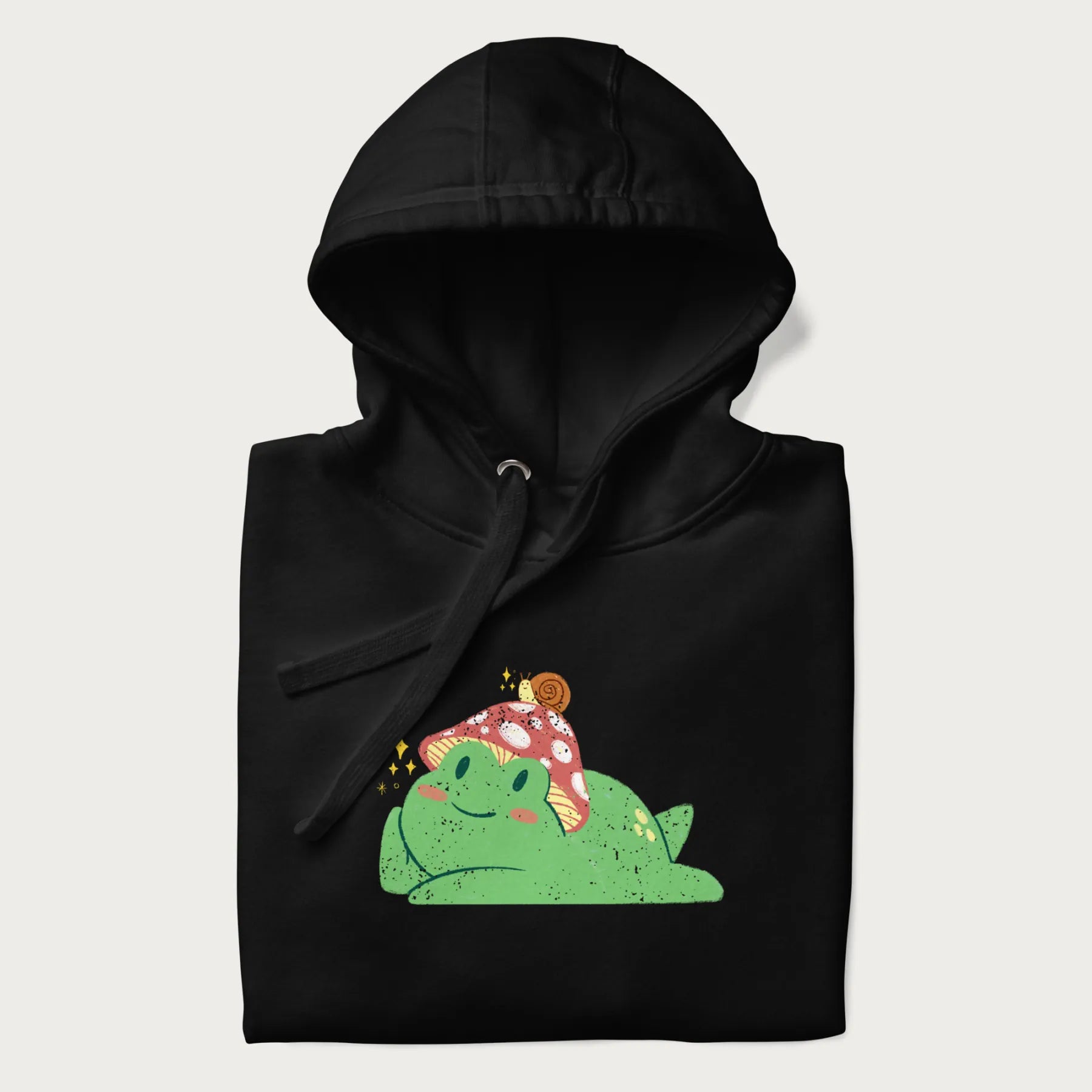 Folded black hoodie with a cottagecore design of a green frog wearing a mushroom cap and a snail on top.