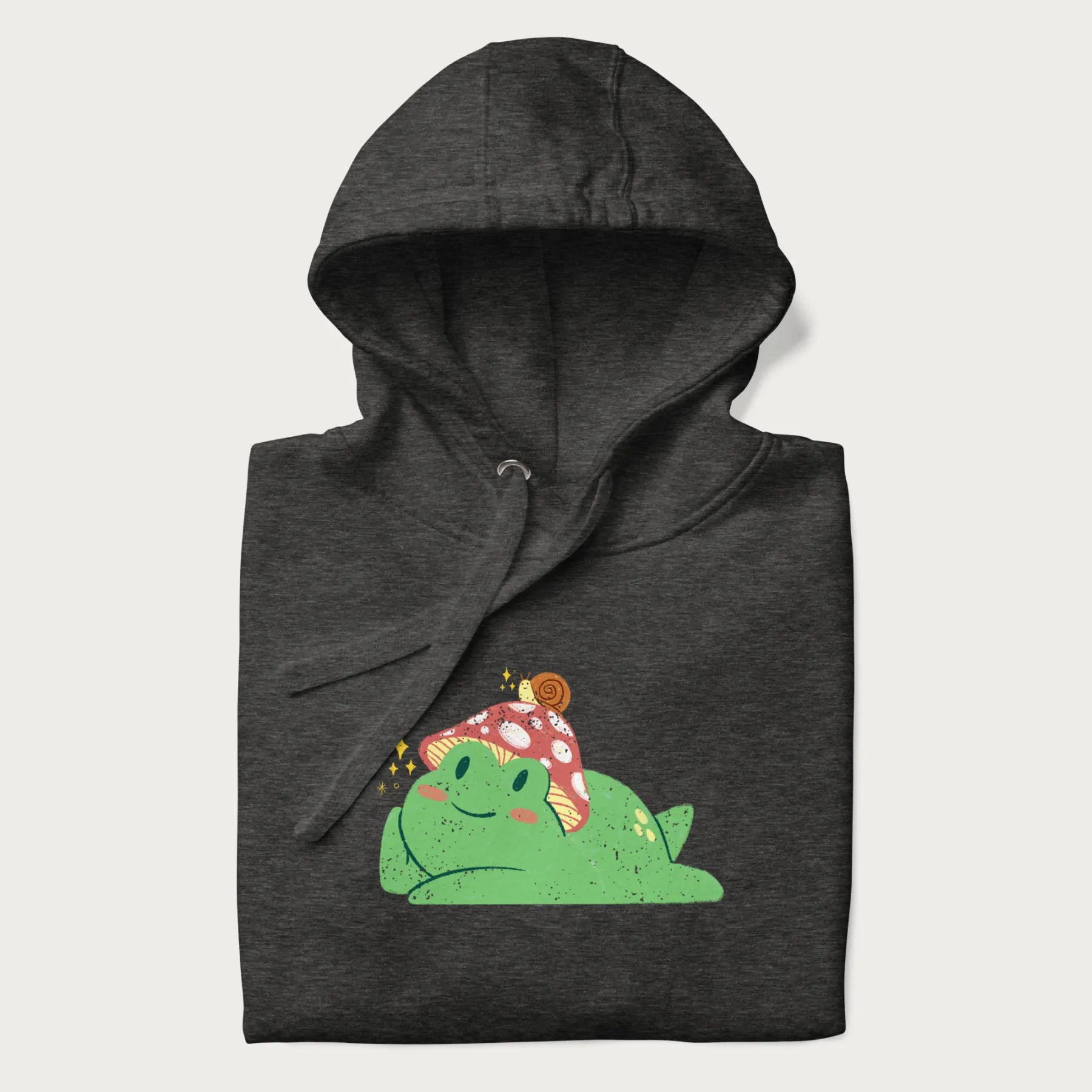 Folded dark grey hoodie with a cottagecore design of a green frog wearing a mushroom cap and a snail on top.